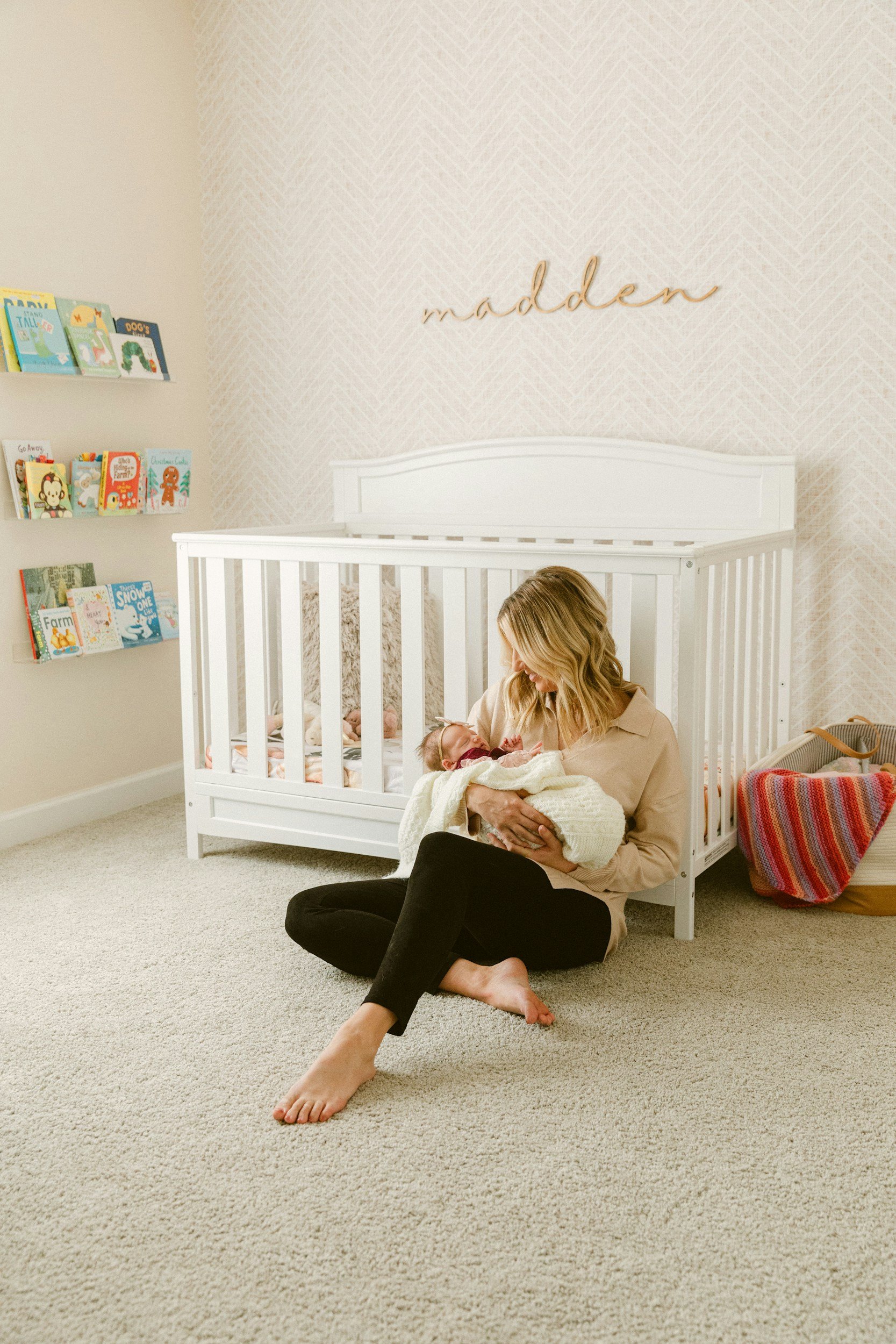 A woman sitting on a beige carpeted floor holding a newborn baby wrapped in a white blanket in a nursery with a white crib, books on a wall shelf, and a colorful bag.