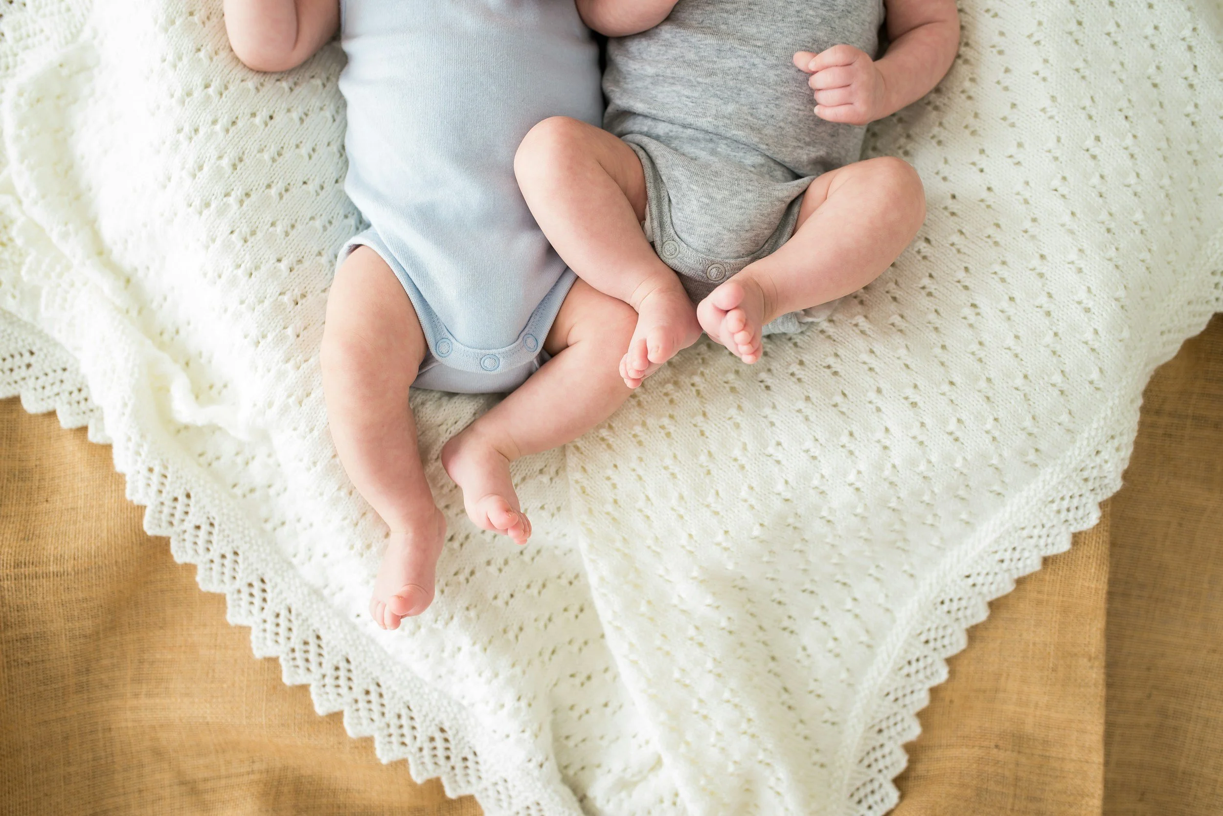Close-up of two babies lying on a cream-colored crocheted blanket on a wooden surface, showing their legs and torsos.