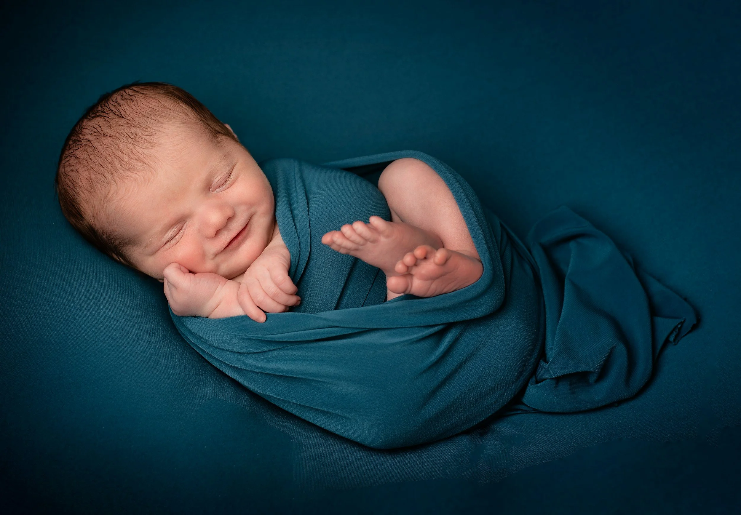 A sleeping newborn baby wrapped in a teal blanket, lying on a teal surface, with a peaceful expression and a slight smile.
