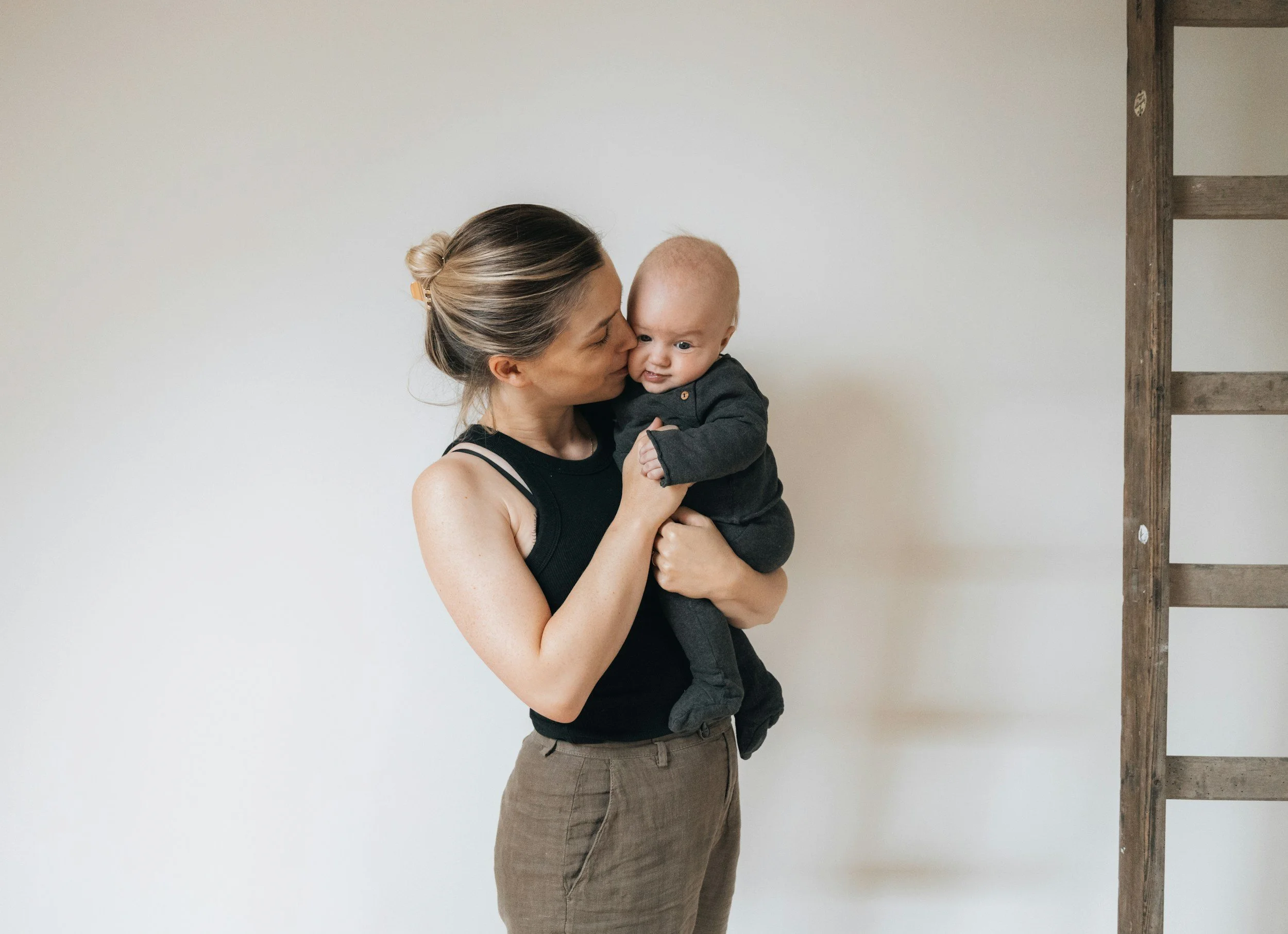 A woman with light brown hair in a bun holding a baby in a black outfit, standing against a plain light-colored wall with a wooden ladder on the right side.