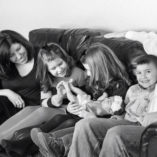 Family of four with a newborn, sitting on a couch, smiling and holding the baby's hand.