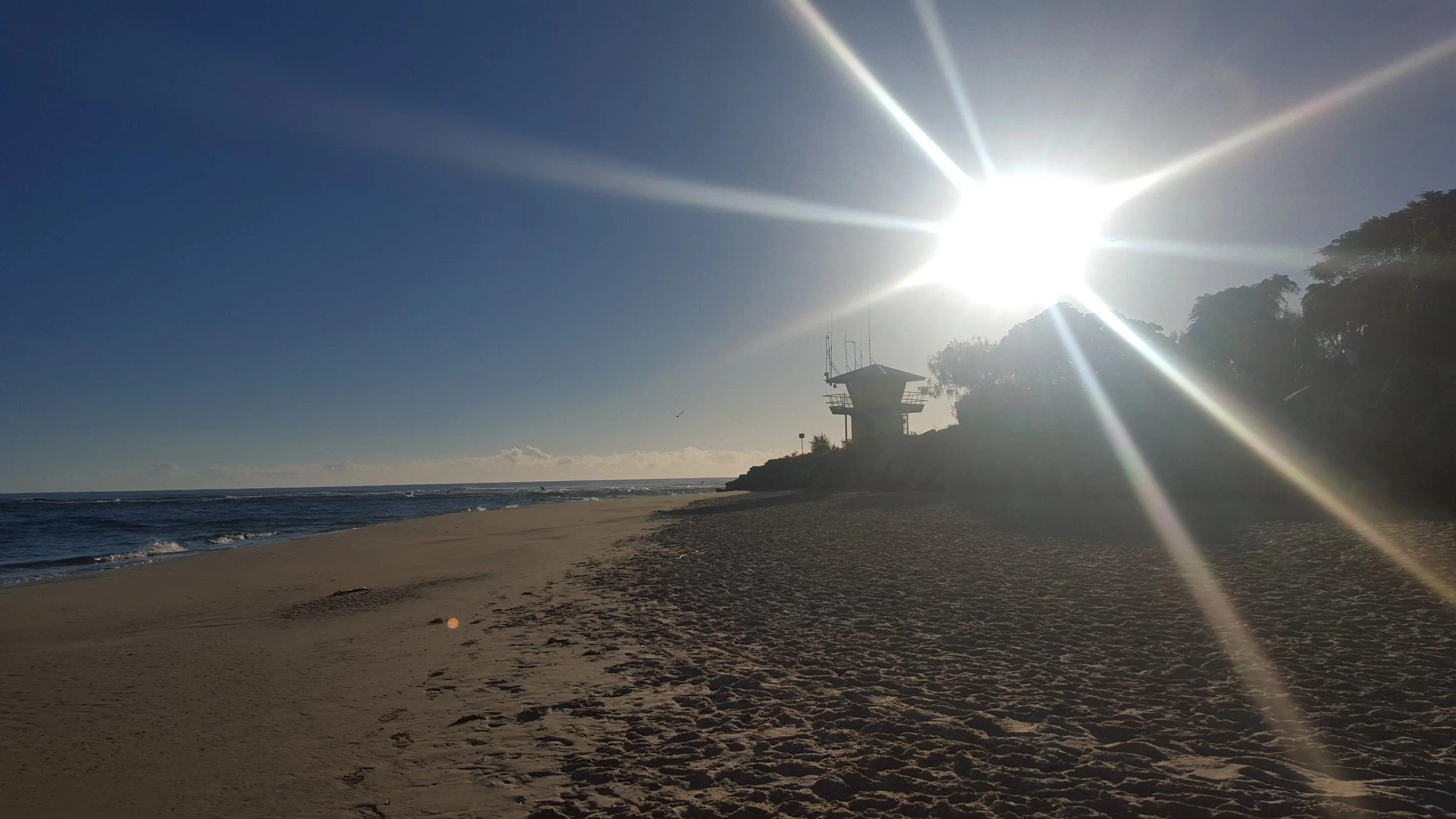 Sunny beach with footprints in the sand, a lifeguard tower, and trees with the bright sun shining overhead.