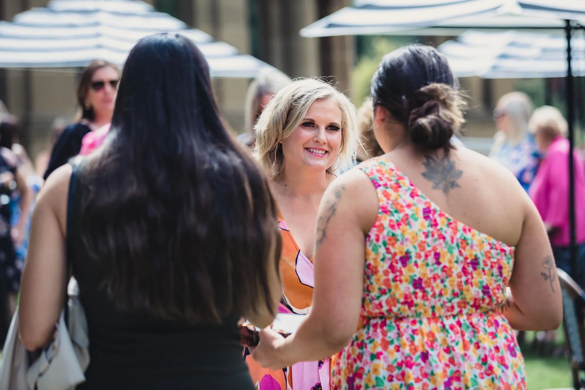 Three women talking outdoors at a social gathering with umbrellas overhead, in colorful summer dresses. The woman in the middle has blonde hair, is smiling, and facing the camera.