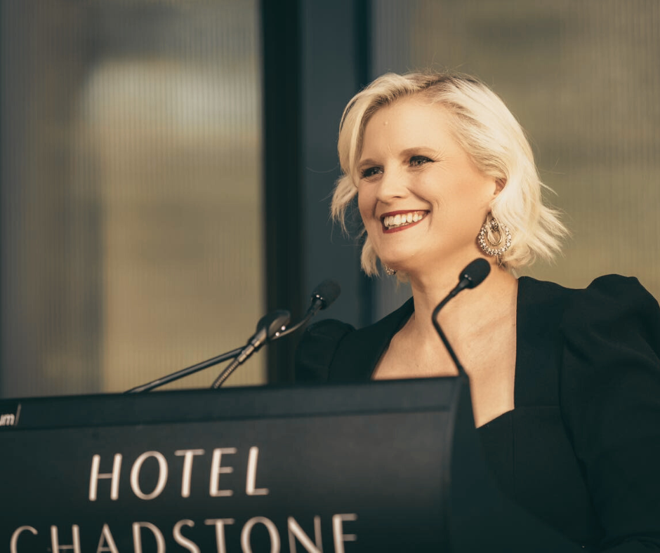A woman with short blond hair wearing earrings and a black outfit standing at a podium labeled 'Hotel Chadstone,' smiling at an event.