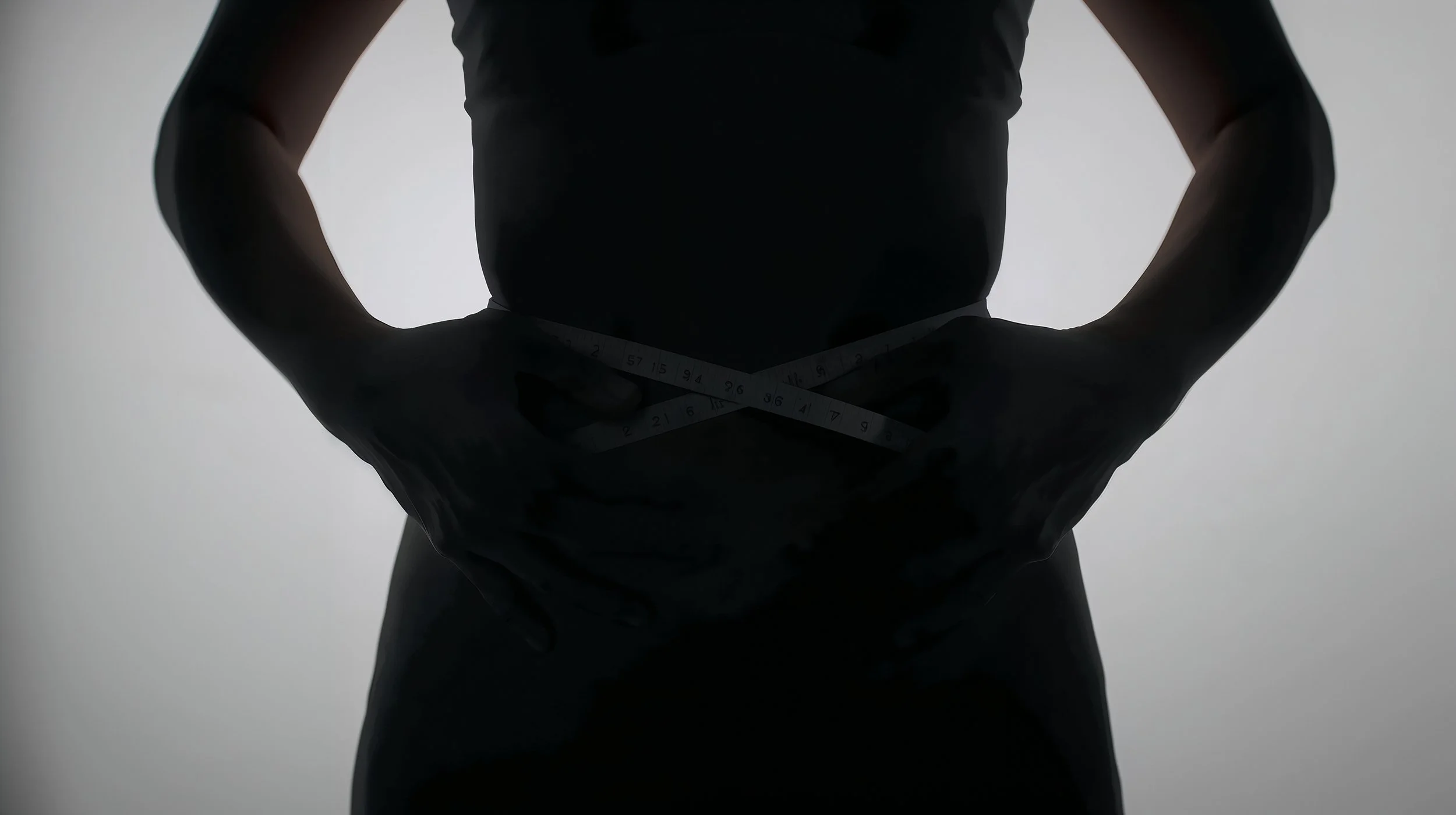 Person in black clothing holding a measuring tape around their waist, backlit with a plain light background.