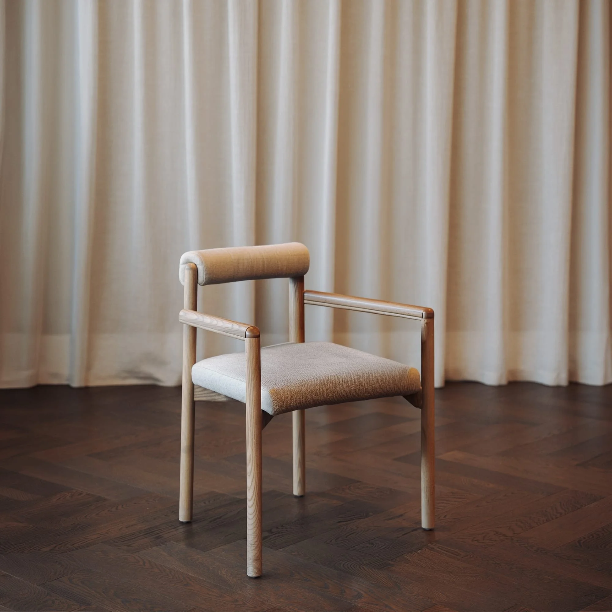 Minimalist wooden armchair with beige cushion, placed in front of cream curtains on a dark wooden floor.