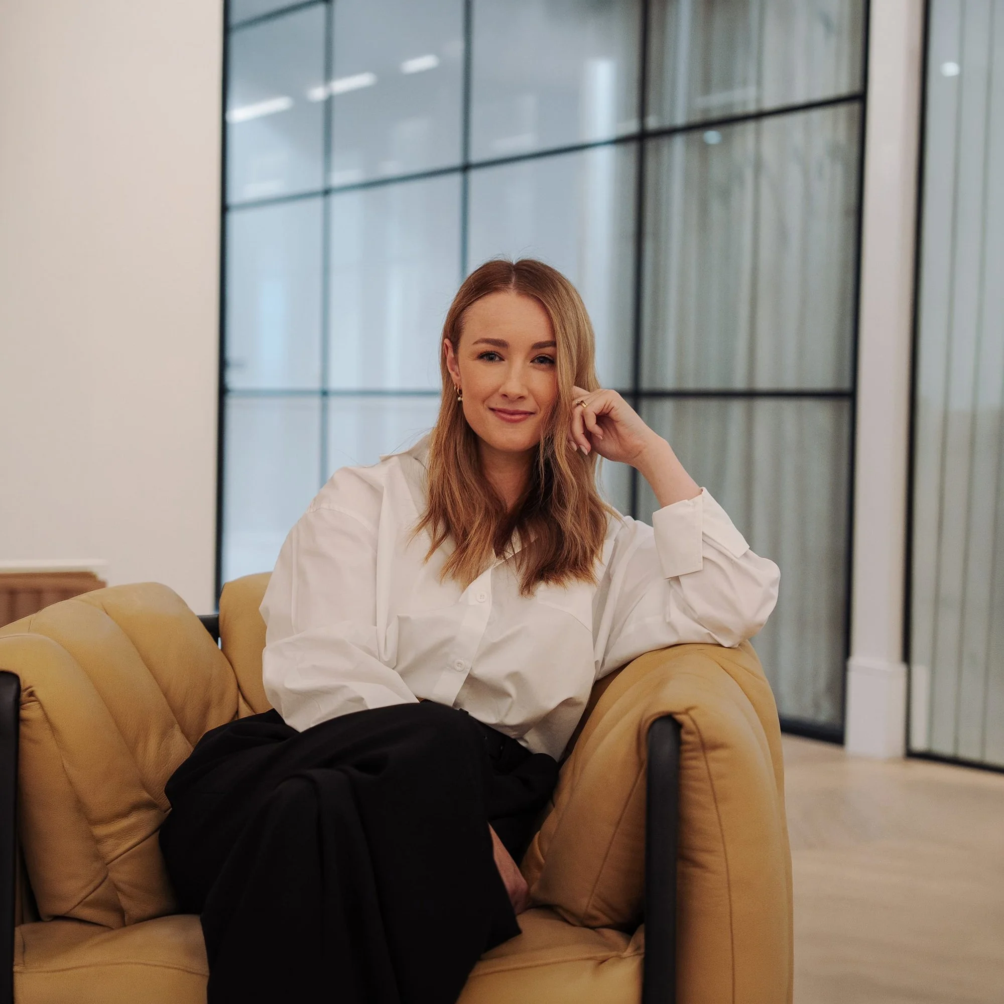 Speaker and leadership coach seated on a couch in a modern office setting, smiling in a relaxed professional environment aligned with communication and leadership development