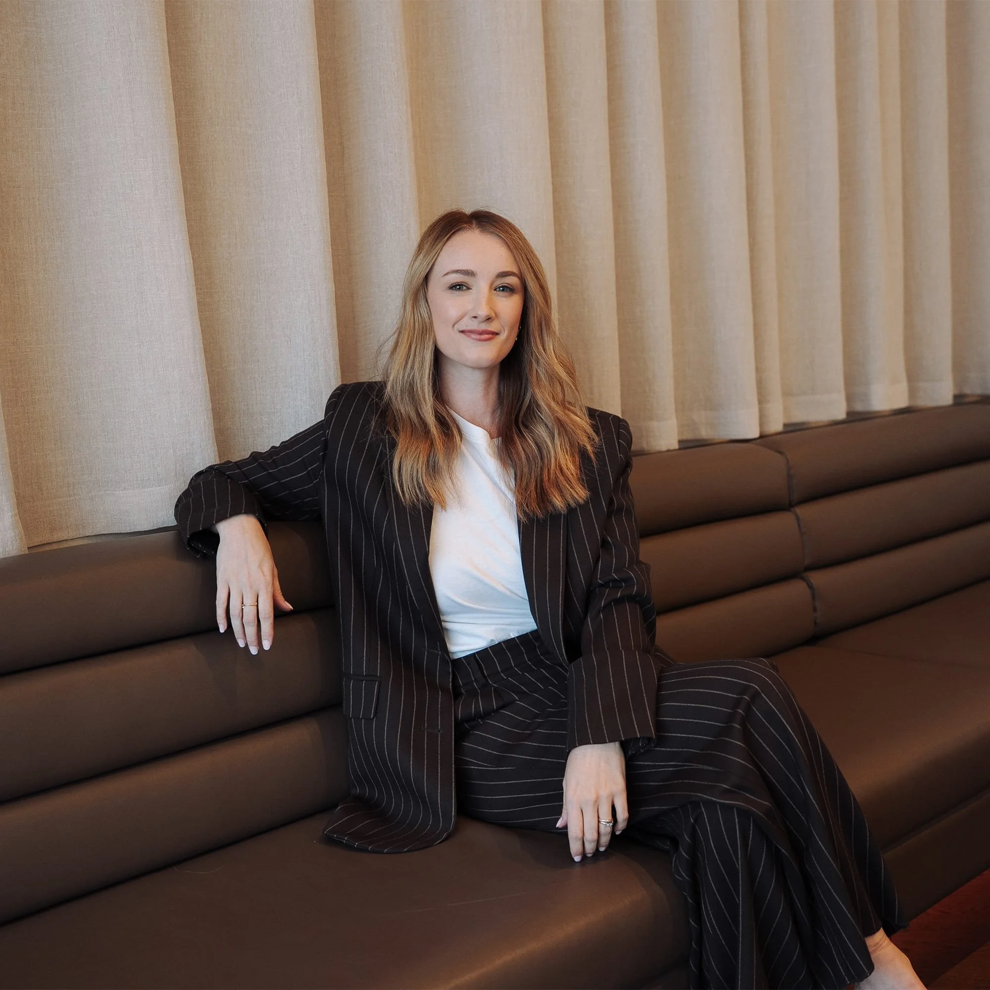 Speaker and facilitator seated on a couch, smiling confidently in a relaxed professional setting, representing leadership coaching and communication training