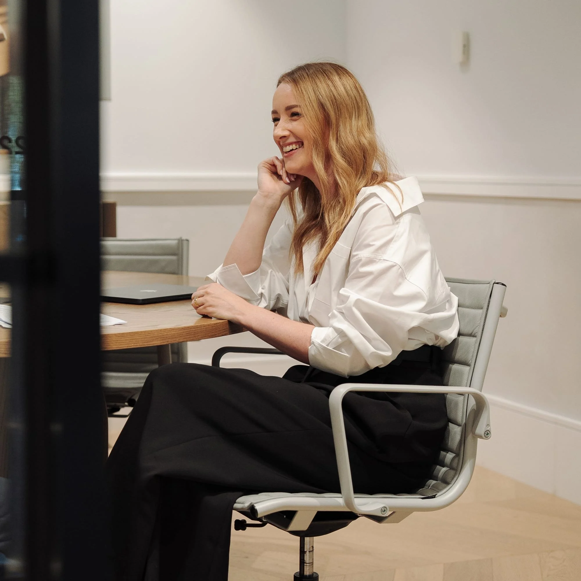Leadership coach and facilitator seated at a conference table, smiling during a team workshop or professional development session with a laptop in front of her