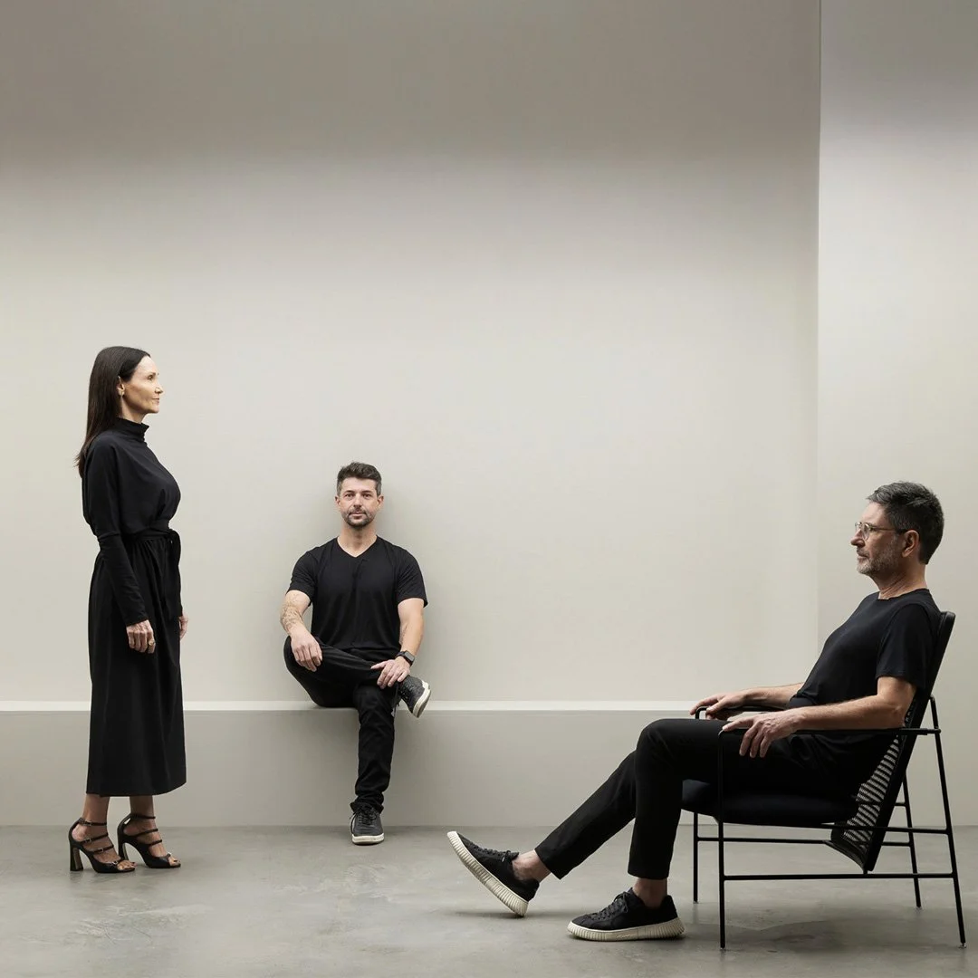 Mulher de vestido preto de salto alto diante de dois homens, um sentado na parede e outro sentado em uma cadeira, em uma sala de paredes claras e piso de concreto.