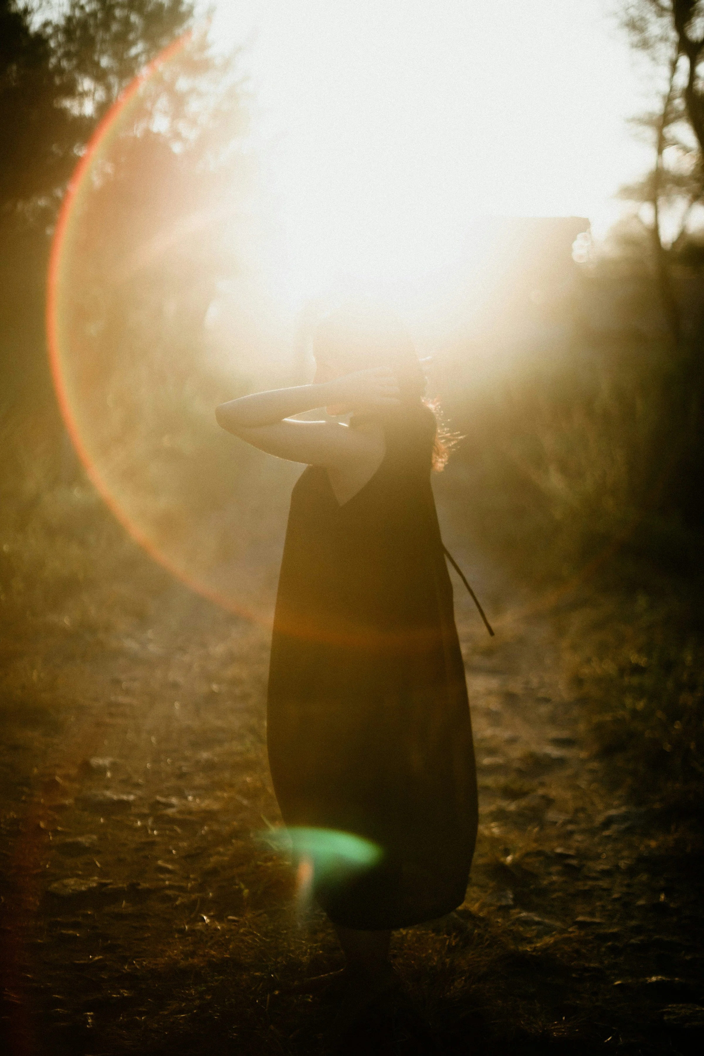 A woman standing outdoors during sunset, with the sun creating a bright glow behind her and a lens flare effect.