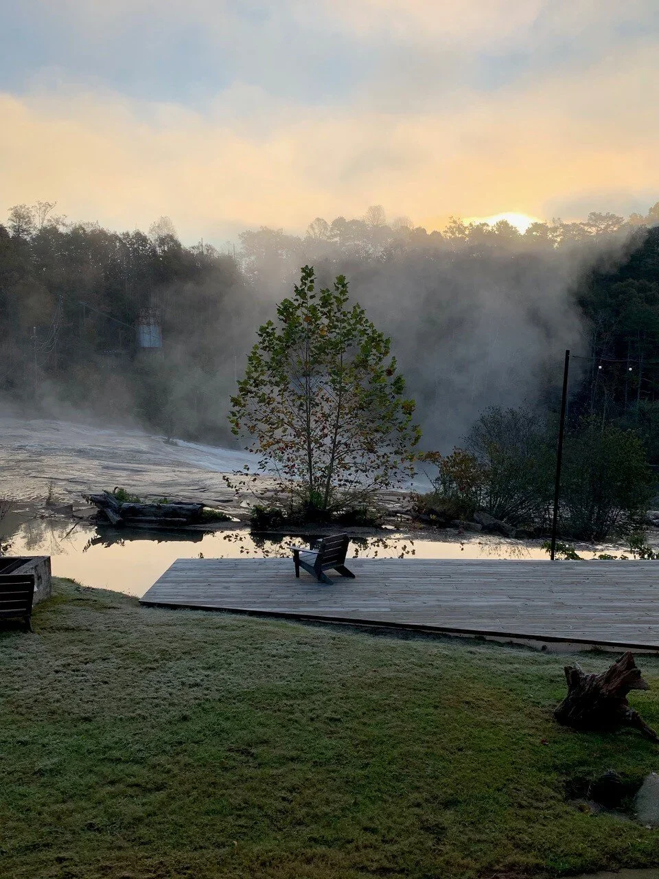 A scenic view of a lake or river with steam rising, surrounded by trees and hills in the background, with a wooden deck and bench in the foreground on a grassy area.