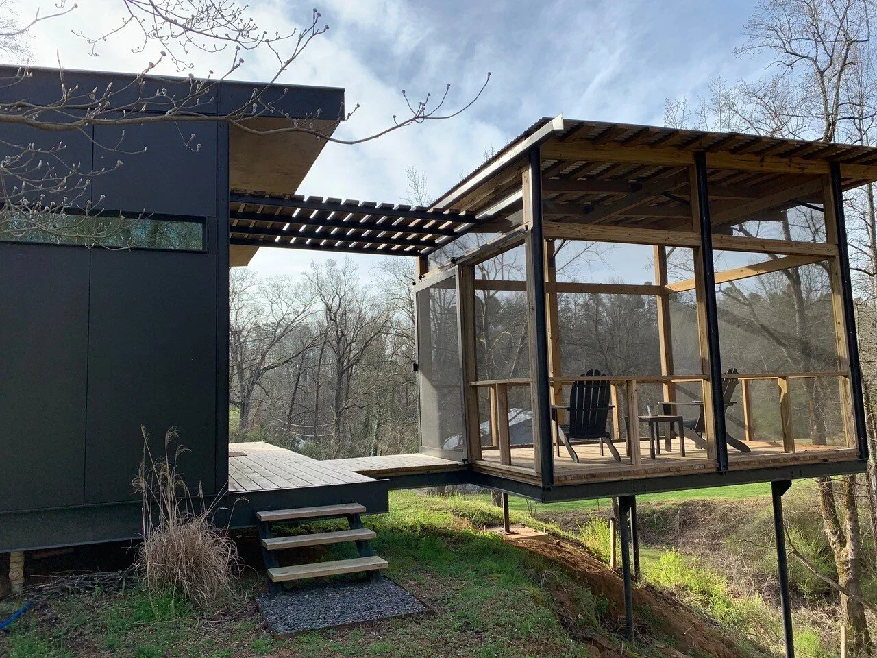Modern house with a black exterior and a screened porch with wooden railings and furniture, situated in a natural landscape with trees and grass.