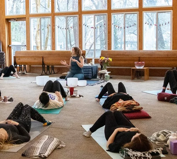 A woman leading a meditation class in a bright room with large windows, seated on the floor with people lying down on mats.