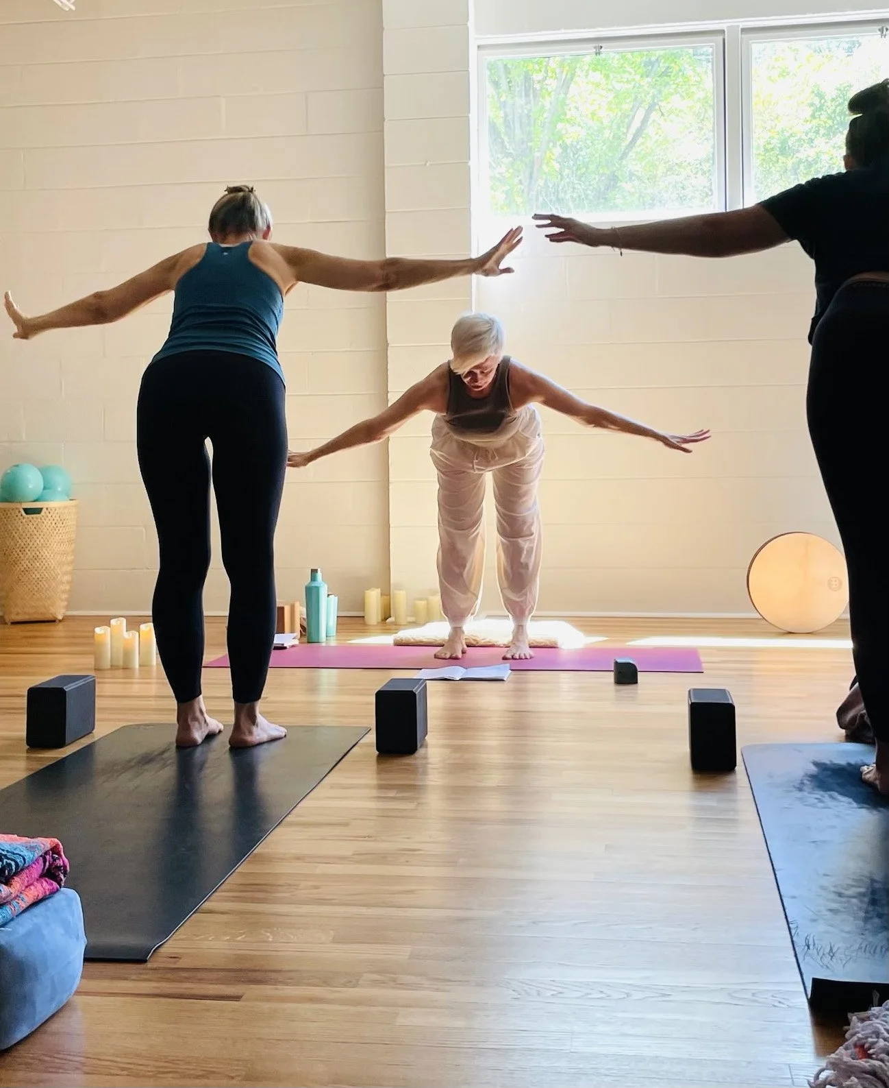 Three women practicing yoga together in a well-lit room with large windows, yoga mats, candles, and exercise props.
