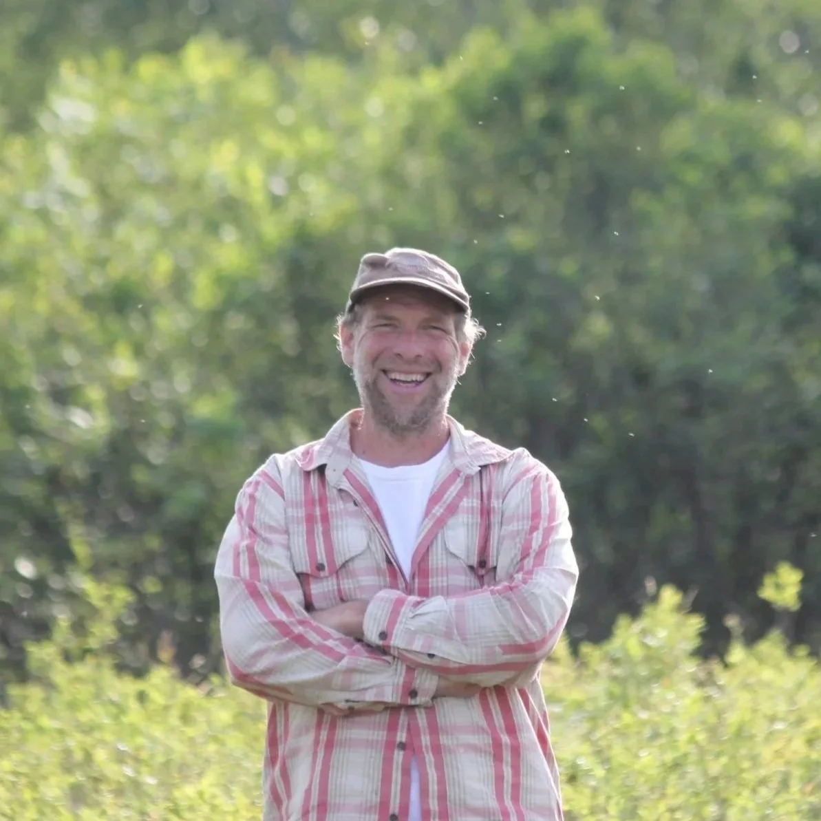 A man standing outdoors with arms crossed, smiling, wearing a plaid shirt and cap, with greenery in the background.