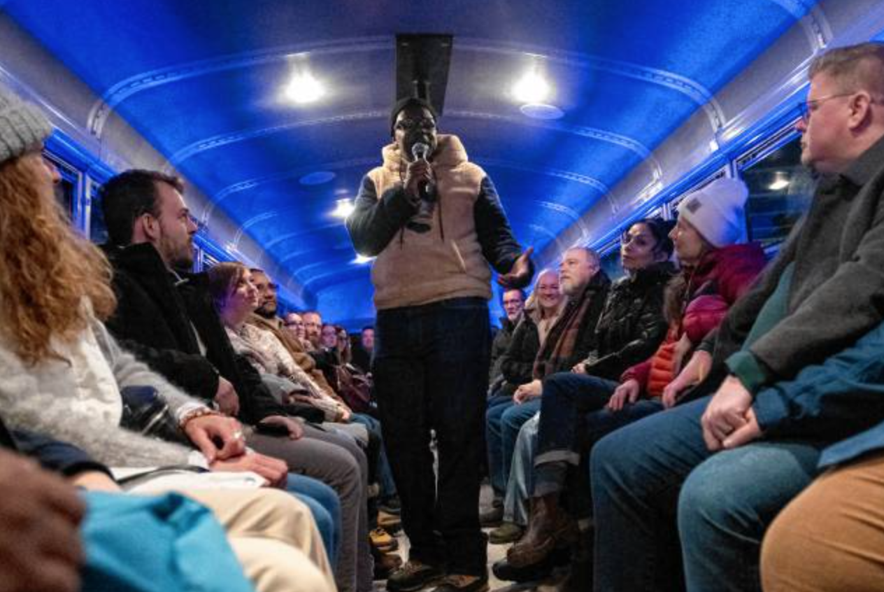 A man standing with a microphone speaking to an audience seated in a a bus with blue lighting.