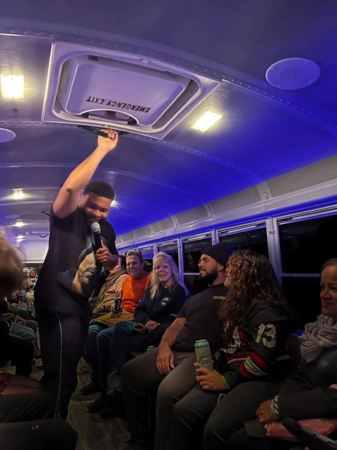 A group of people on a bus, some smiling, some holding drinks, with a man standing holding a microphone and opening an emergency exit hatch on the ceiling.