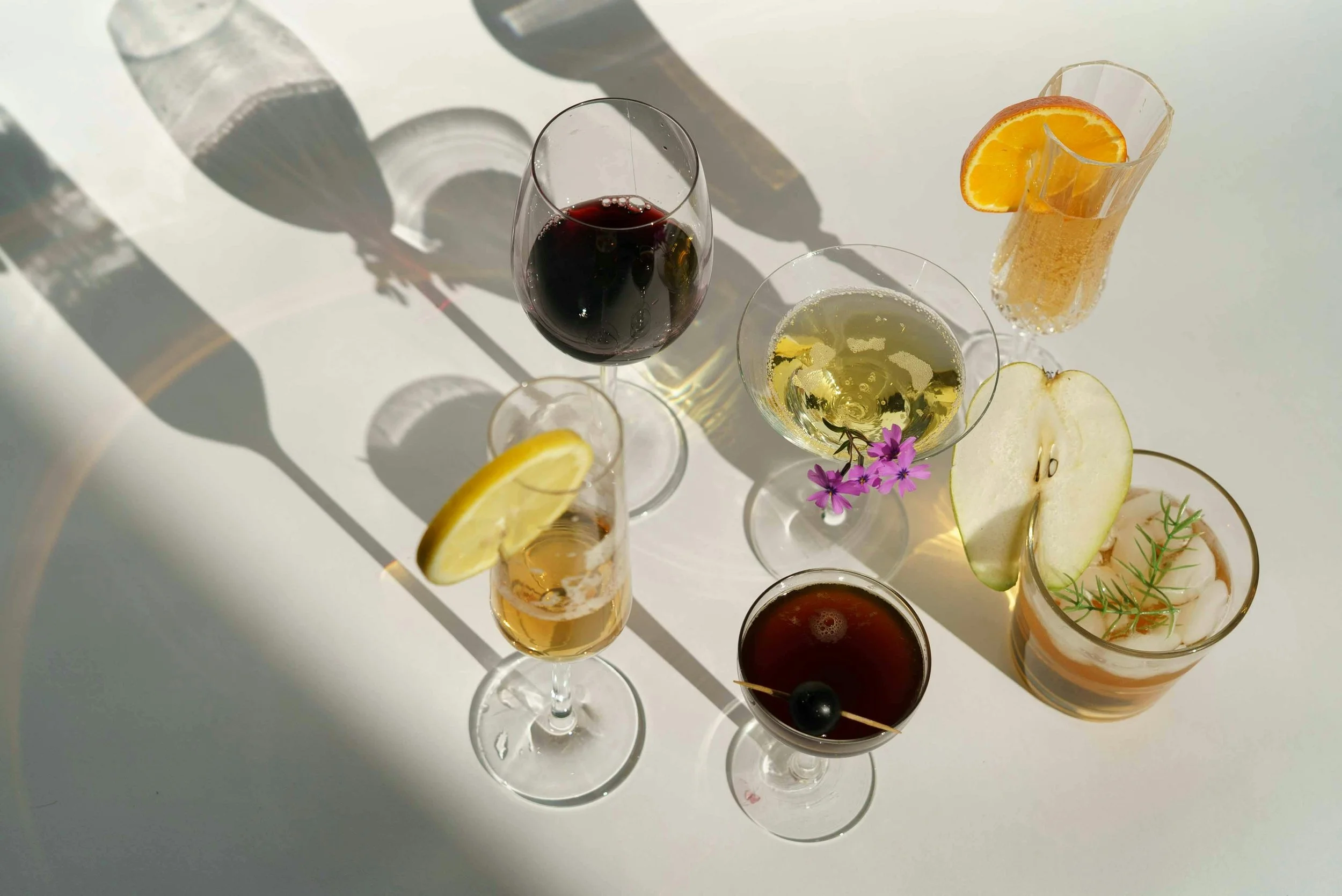 An overhead view of various cocktails on a white surface, with shadows cast by the glasses. The drinks include red wine, champagne with a lemon slice, a drink with an orange slice, and others with garnishes like lemon, apple slices, and herbs.