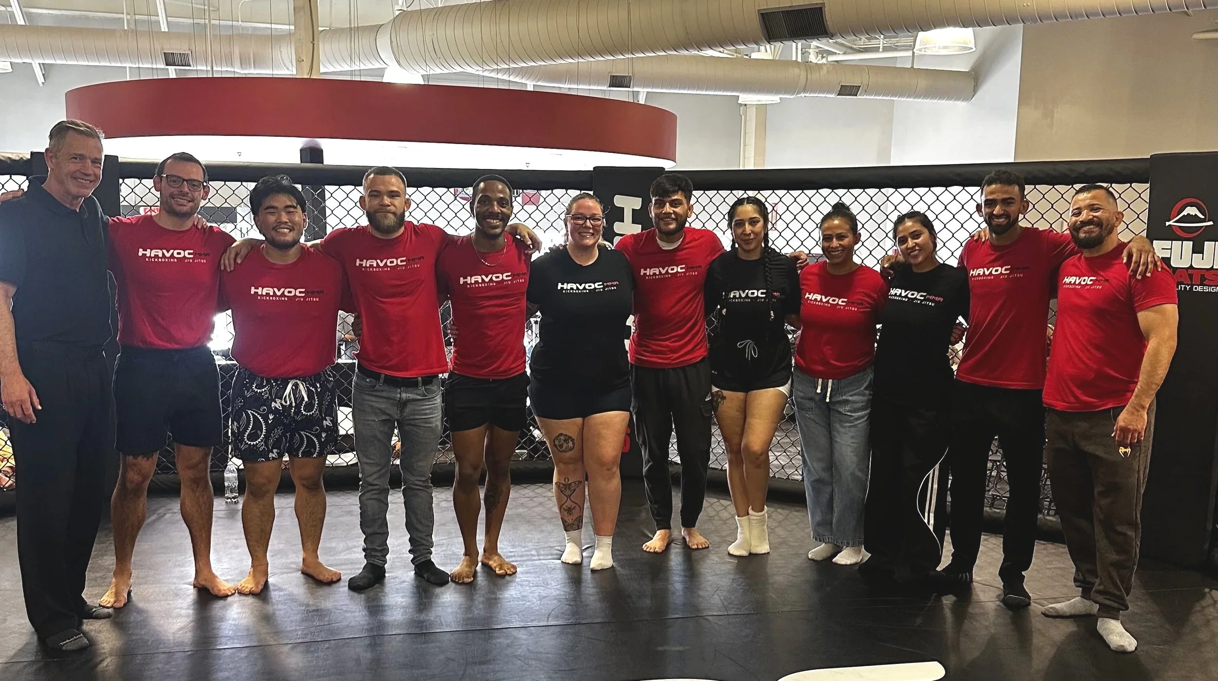 Group of people inside a sports gym, standing in front of a cage, smiling with arms around each other's shoulders. Community of people in martial arts.