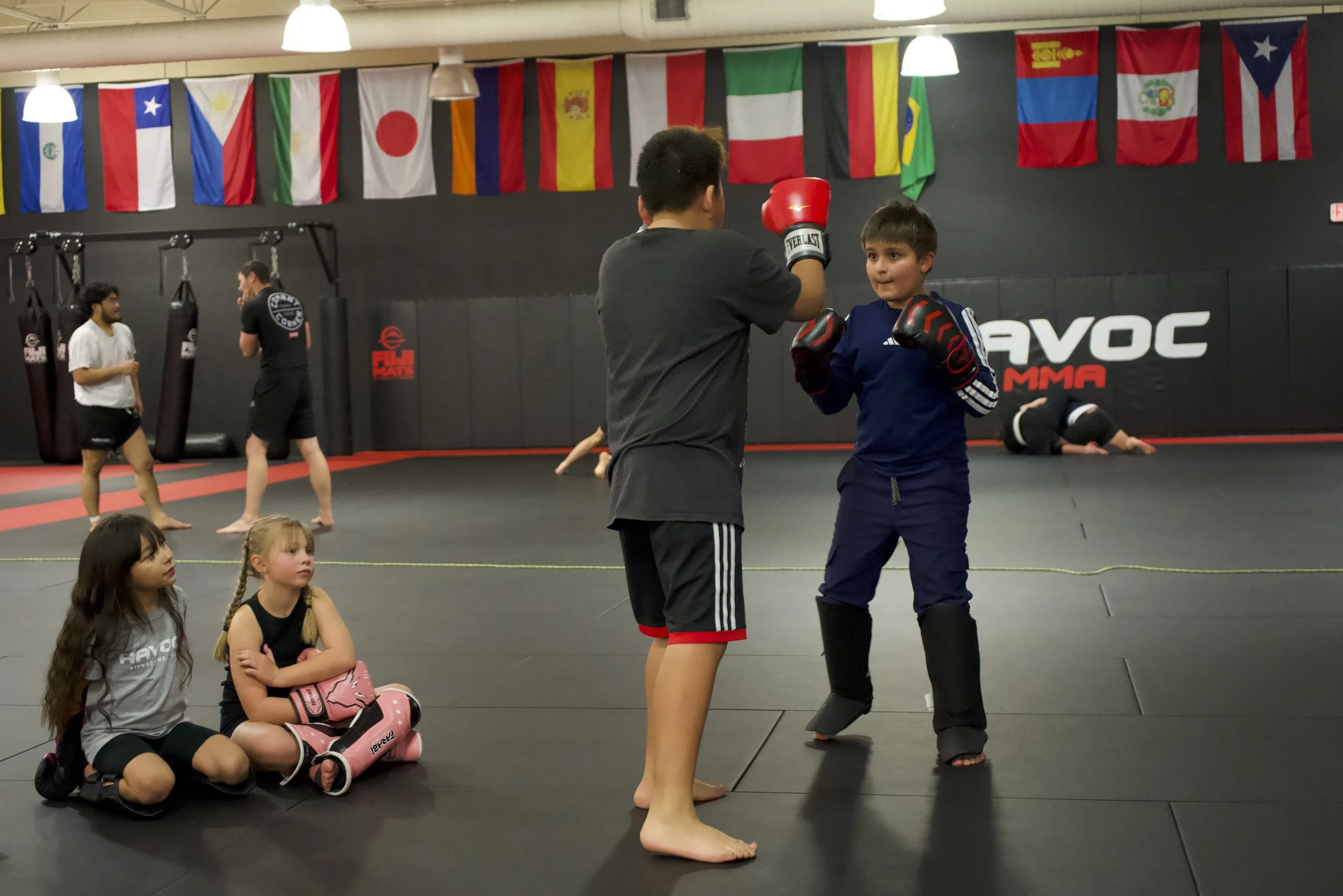 Two children practicing boxing, one is holding focus mitts, and the other is wearing boxing gloves. Two girls are sitting on the floor observing, while others train in the background at a gym with international flags hanging above.