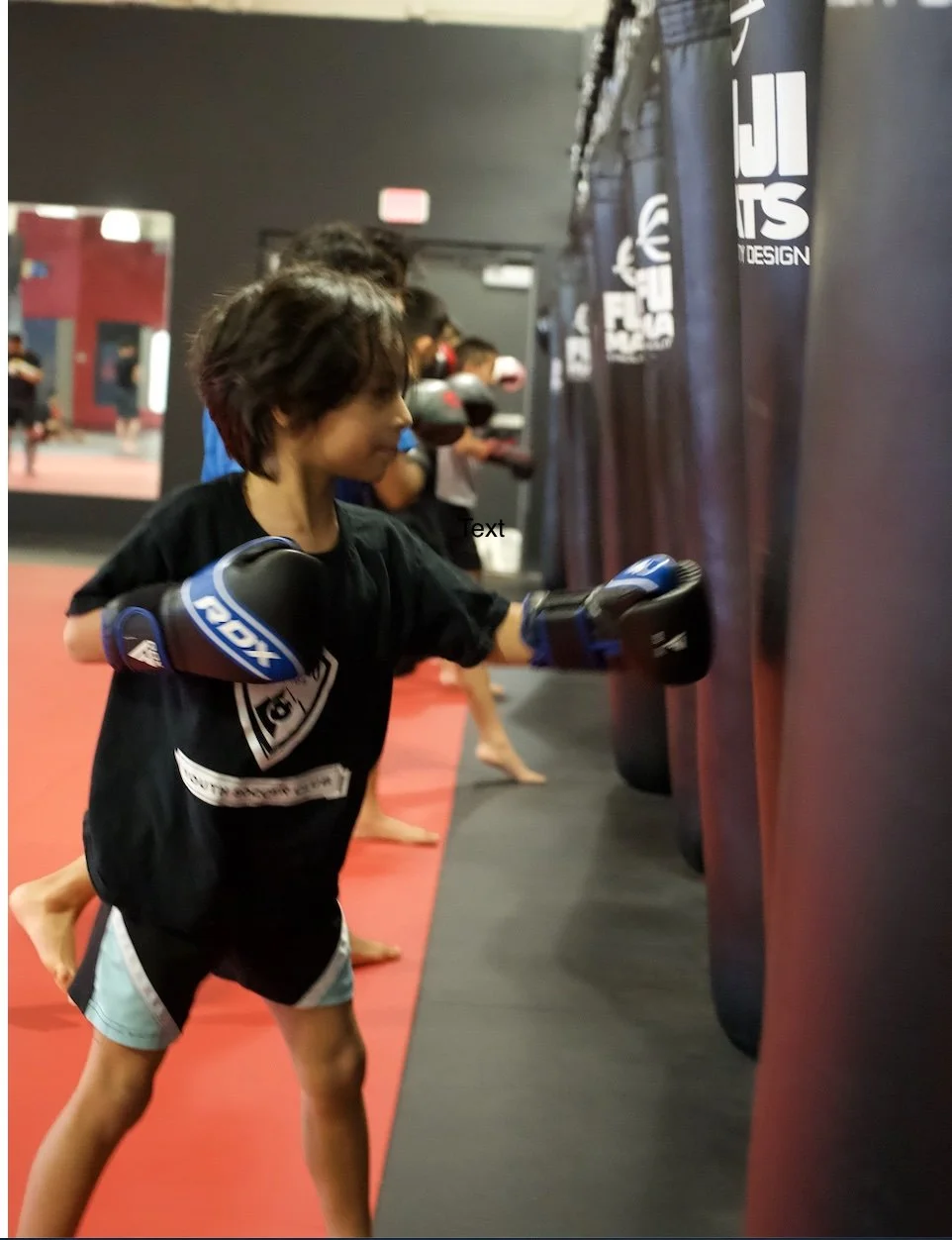 Young girl practicing boxing, wearing gloves, hitting a punching bag in a gym with other kids in the background.