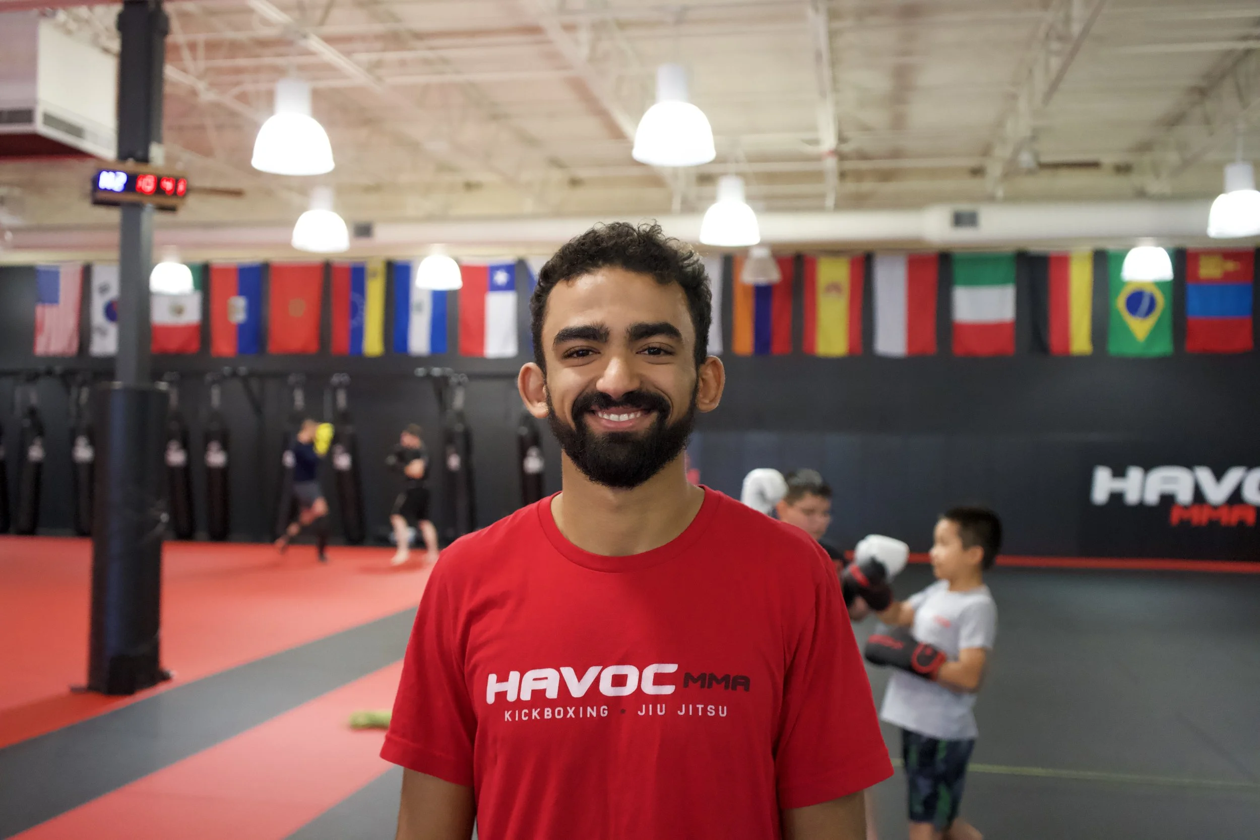 A smiling young man with dark hair and a beard wearing a red T-shirt with 'HAVOC MMA' printed on it, standing inside a martial arts gym with children practicing in the background.