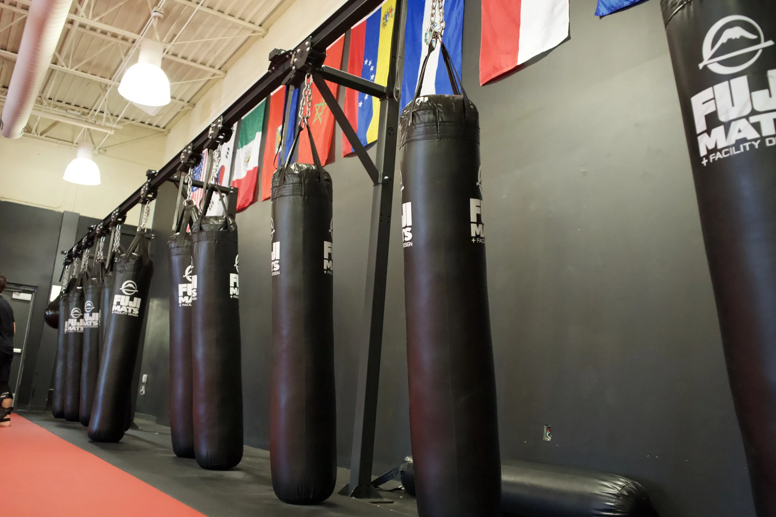 A row of black punching bags hanging from a metal frame inside a martial arts gym, with flags hanging on the wall behind them.