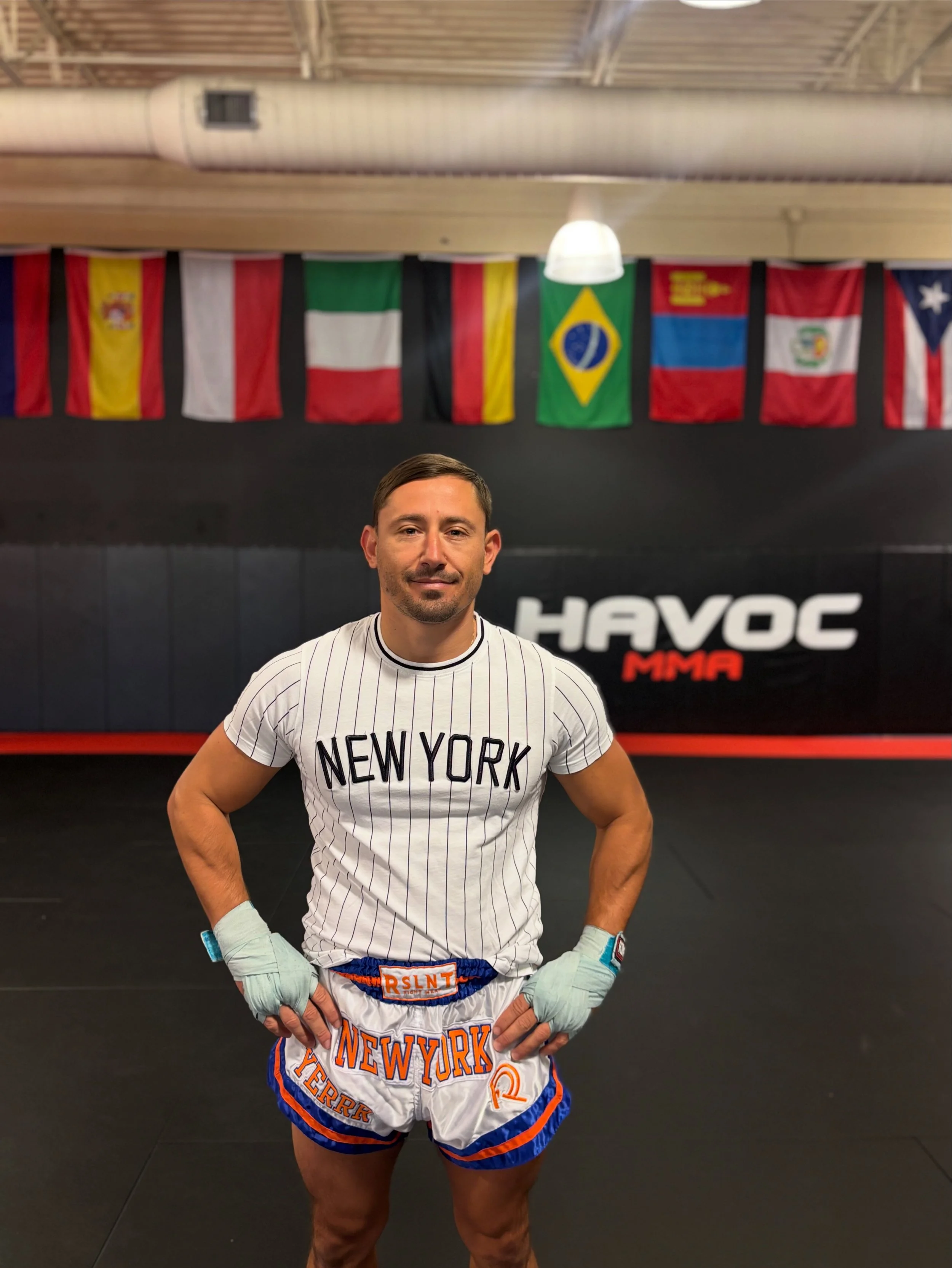 A man in a white New York sports jersey and shorts stands in a gym with boxing gloves, in front of a black and red Havoc MMA sign and international flags hanging overhead.