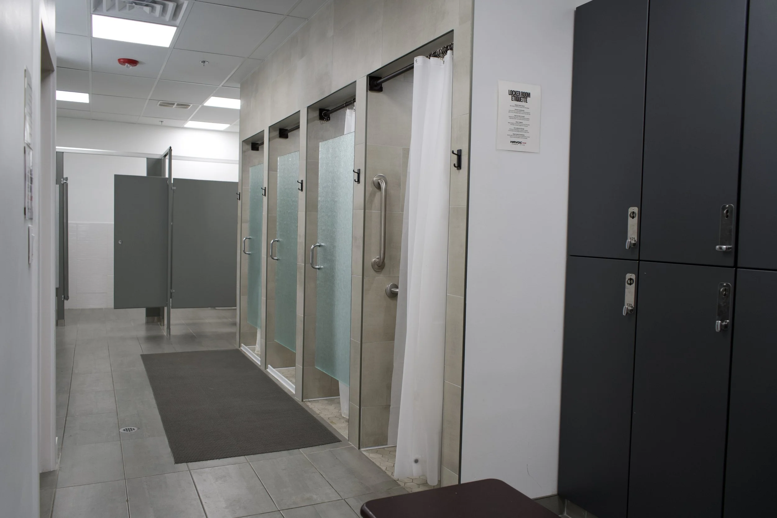 Public restroom with four shower stalls, privacy curtains, and lockers on the right side, in a commercial building.