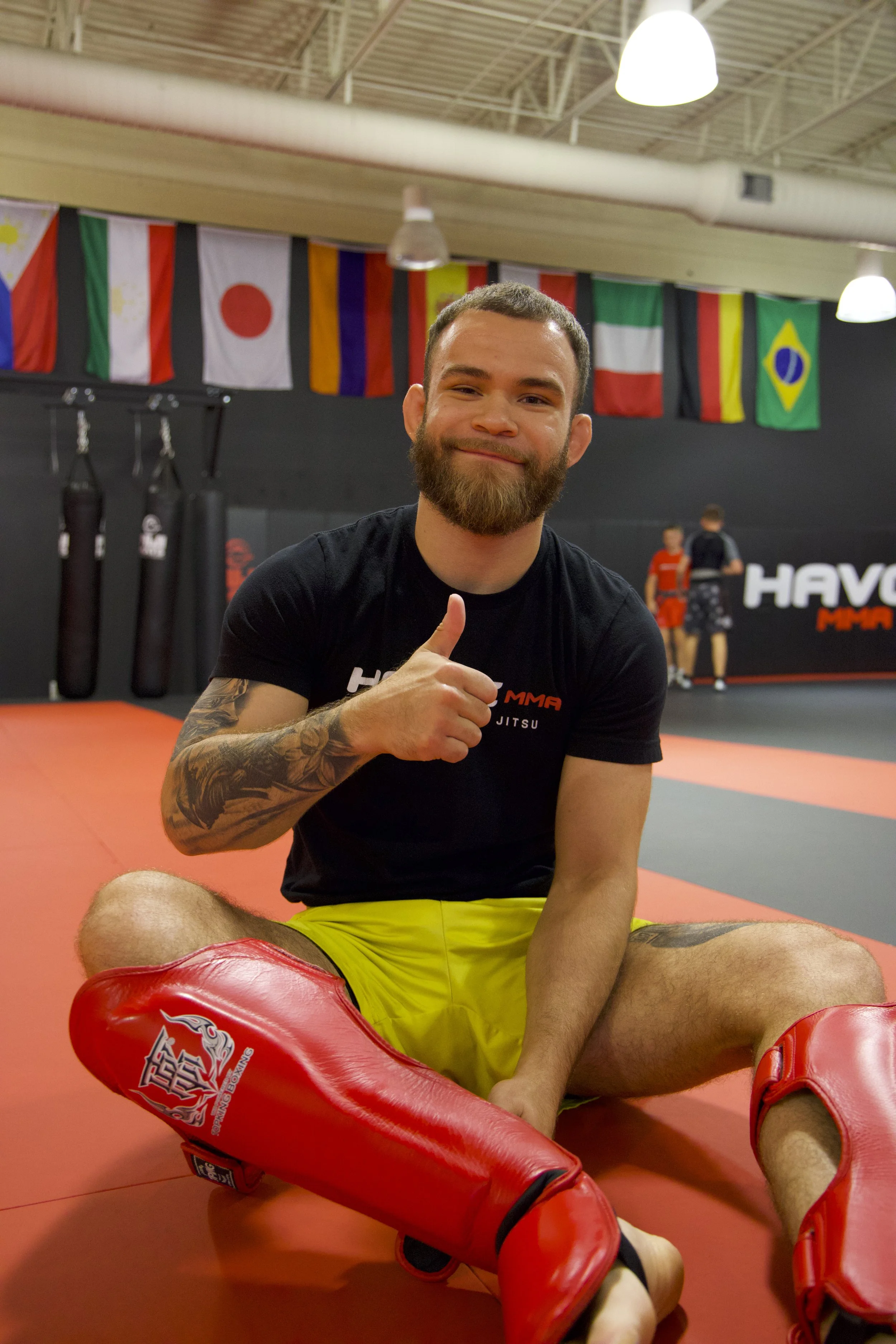 A man with a beard and tattoos sitting cross-legged on a martial arts mat, giving a thumbs-up. He is wearing red shin guards, yellow shorts, and a black t-shirt with red and white lettering. International flags hang from the ceiling in the background, with other people practicing in a martial arts gym.