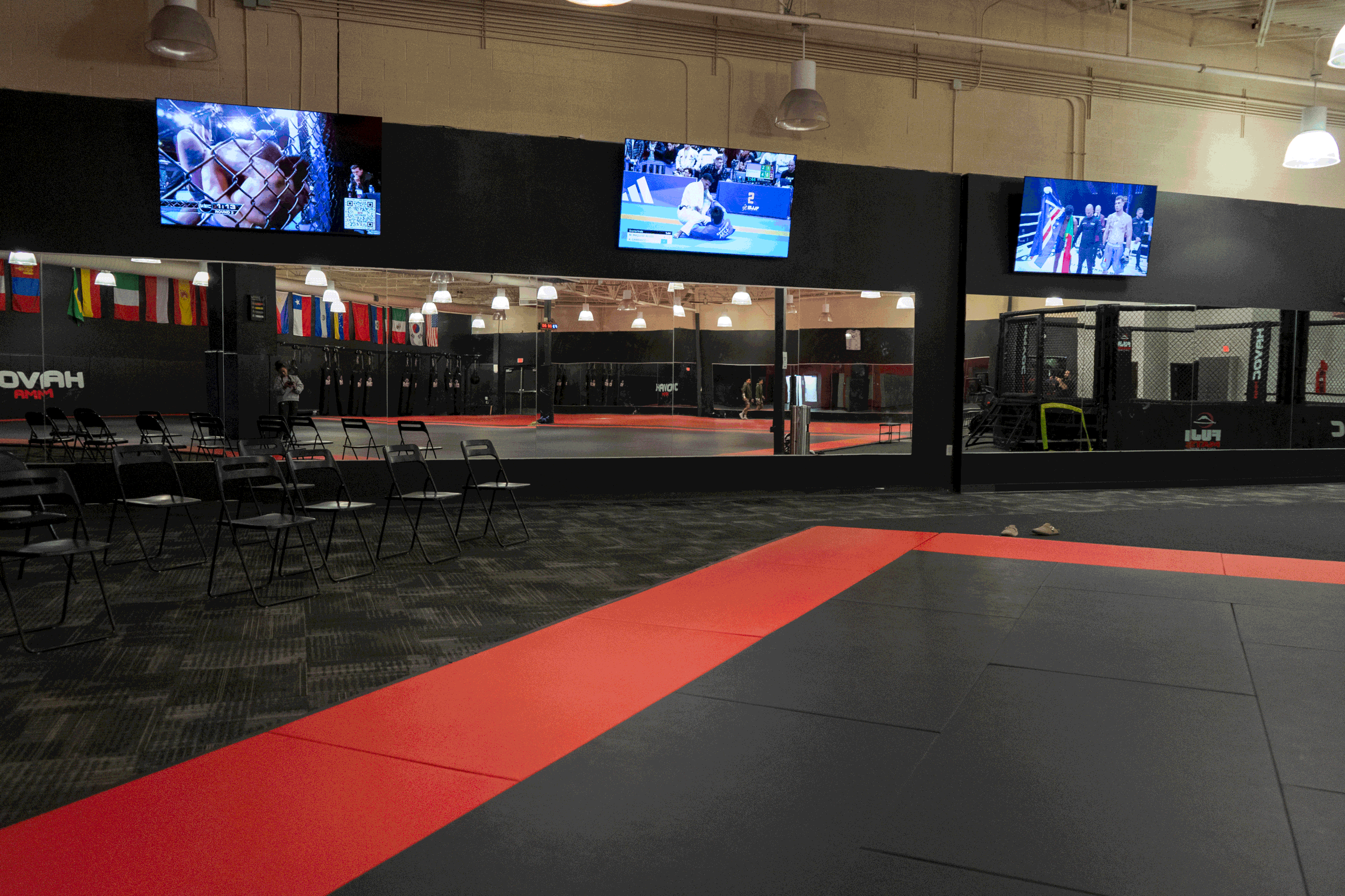 Empty MMA gym with black and red mats, chairs, and three television screens showing combat sports, with international flags on the wall and a mirror reflecting the training area.