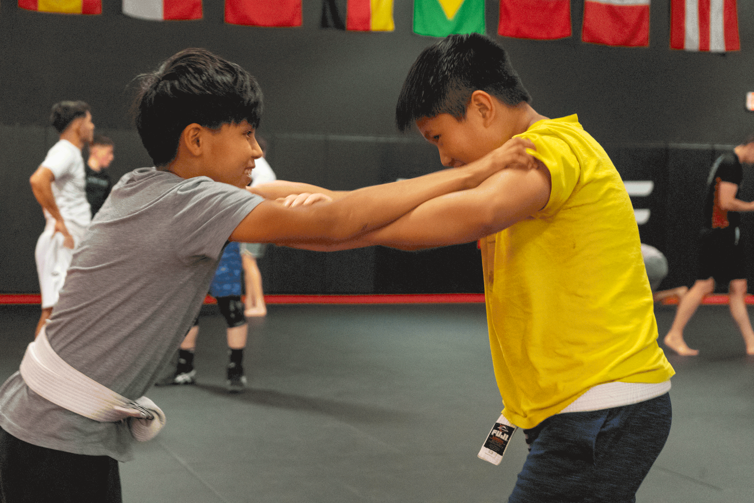 Two boys practicing martial arts in a gym, engaging in a sparring drill with one boy wearing a gray shirt and a white belt, and the other in a yellow shirt. Other children are practicing in the background.