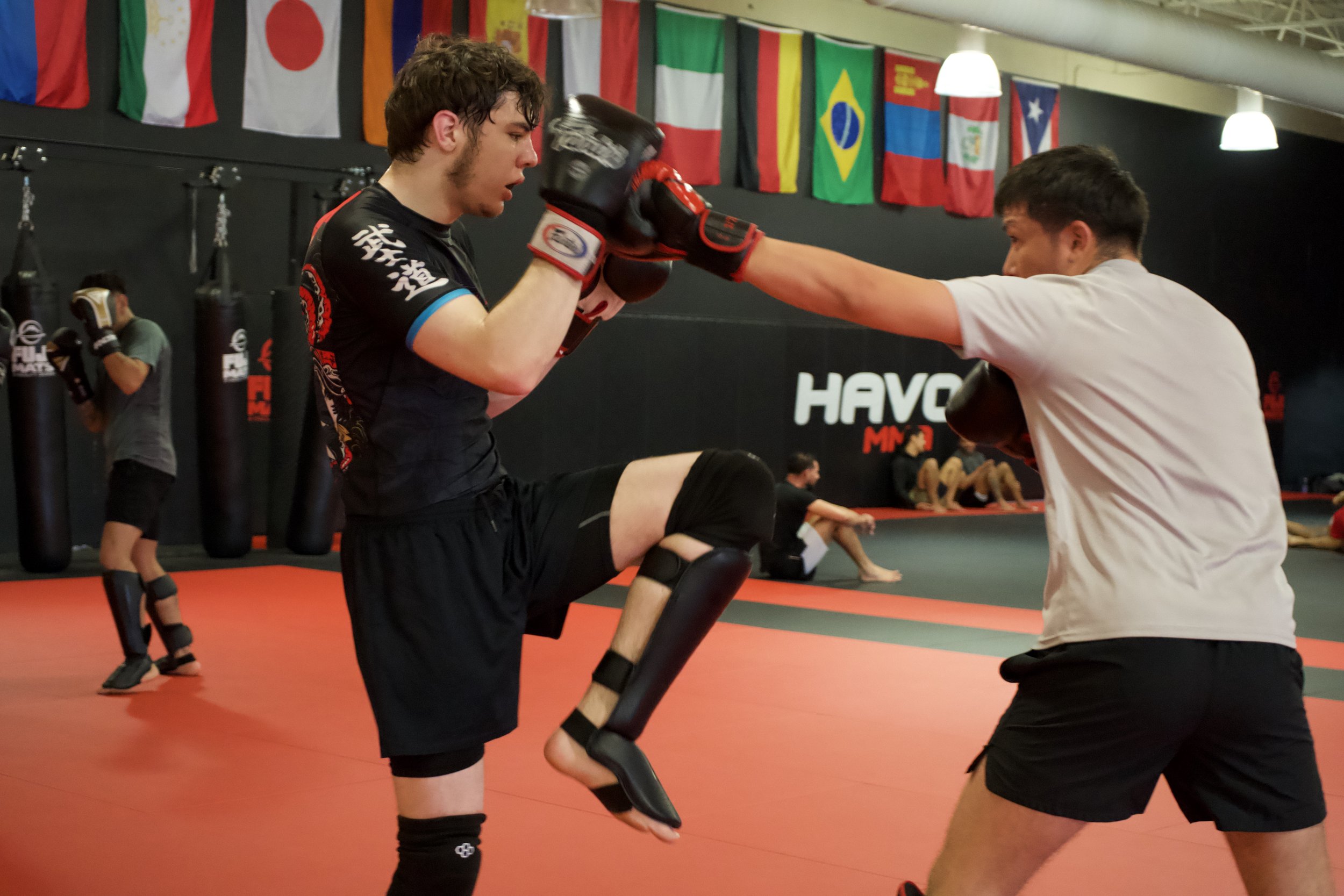 Two people practicing martial arts in a gym. One person is throwing a punch while the other blocks. They wear gloves, shin guards, and athletic clothing. Flags from various countries hang on the wall behind them, and other individuals are seen training in the background.