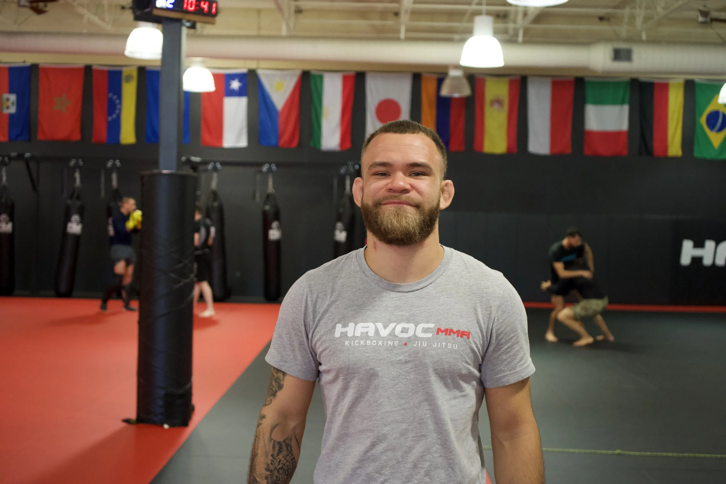 A man with a beard and tattoos wearing a gray 'HAVOC MMA' T-shirt, standing inside a martial arts gym with flags of various countries hanging from the ceiling and people training in the background.