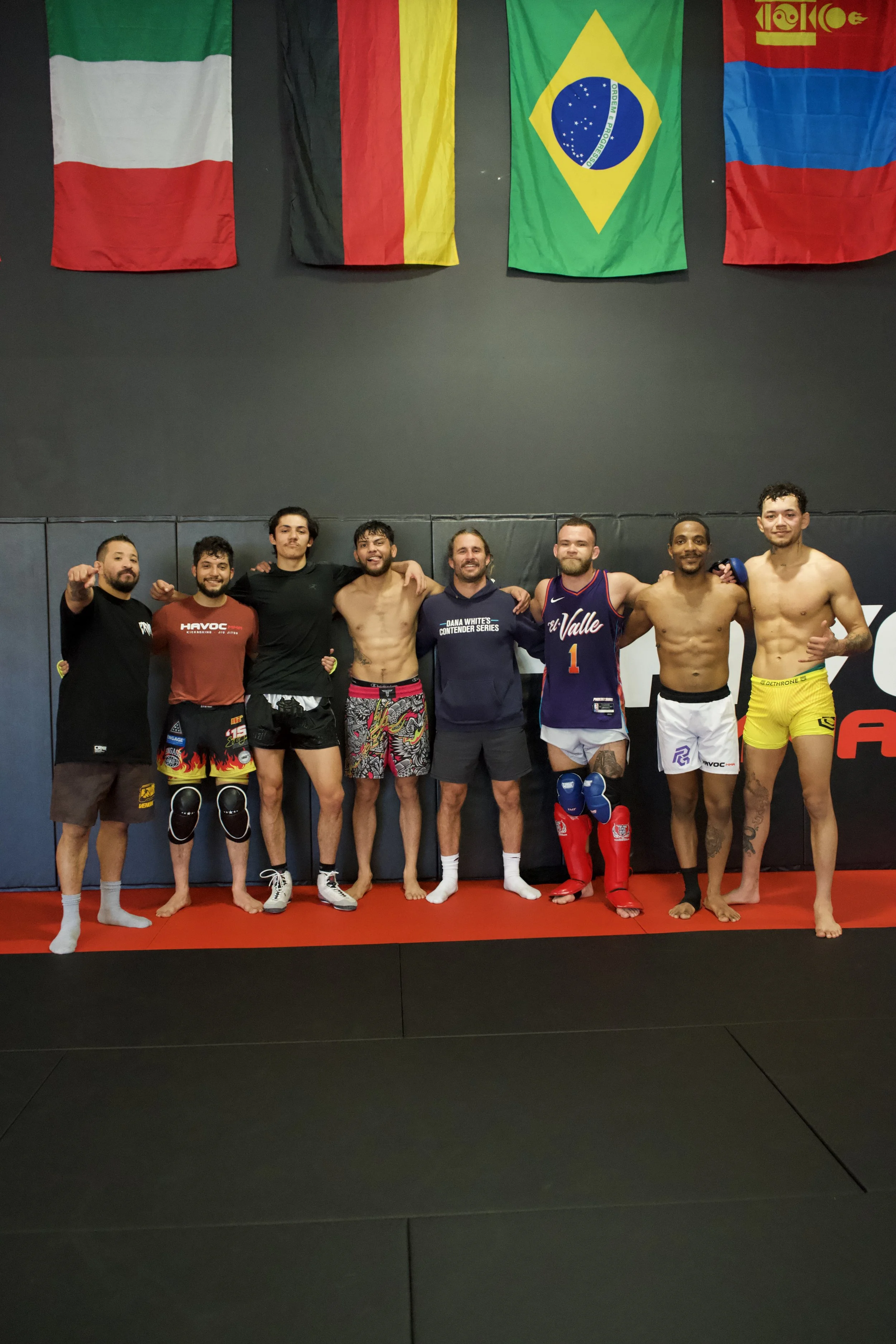 A group of eight men standing together on a martial arts gym floor, smiling with their arms around each other, in front of international flags hanging on the wall.