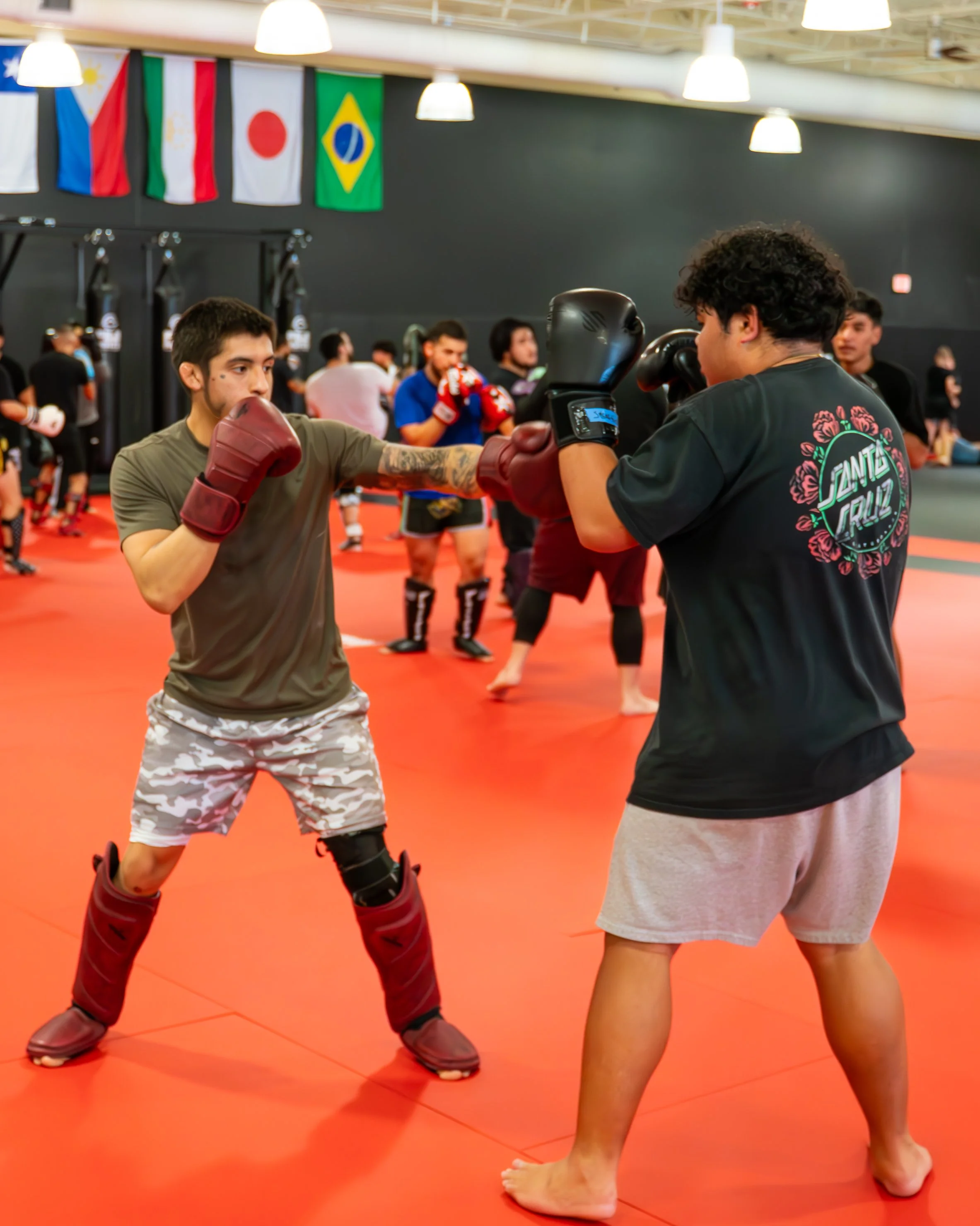 Two people sparring in a martial arts gym, practicing boxing with gloves and mitts, surrounded by other fighters and flags from various countries hanging from the ceiling.
