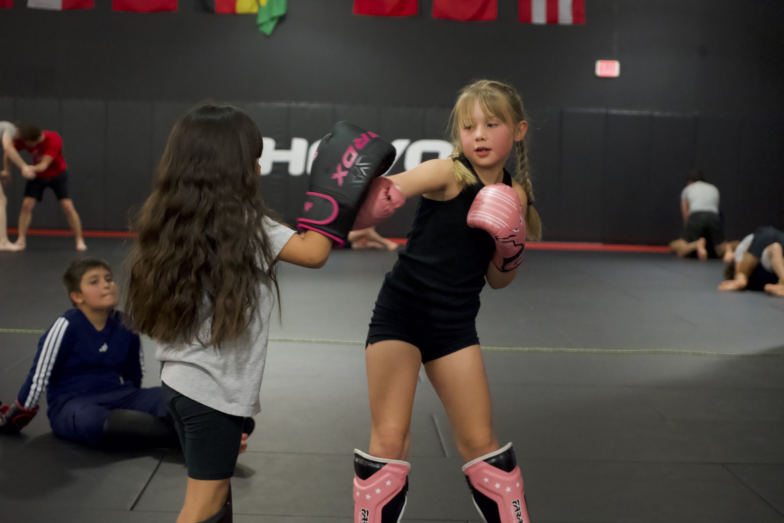 Two young girls practicing boxing in a gym, with one girl wearing pink boxing gloves, pink shin guards, and black workout clothes, while the other girl wears grey shirt, black pants, and pink boxing gloves. The girl in pink is throwing a punch.
