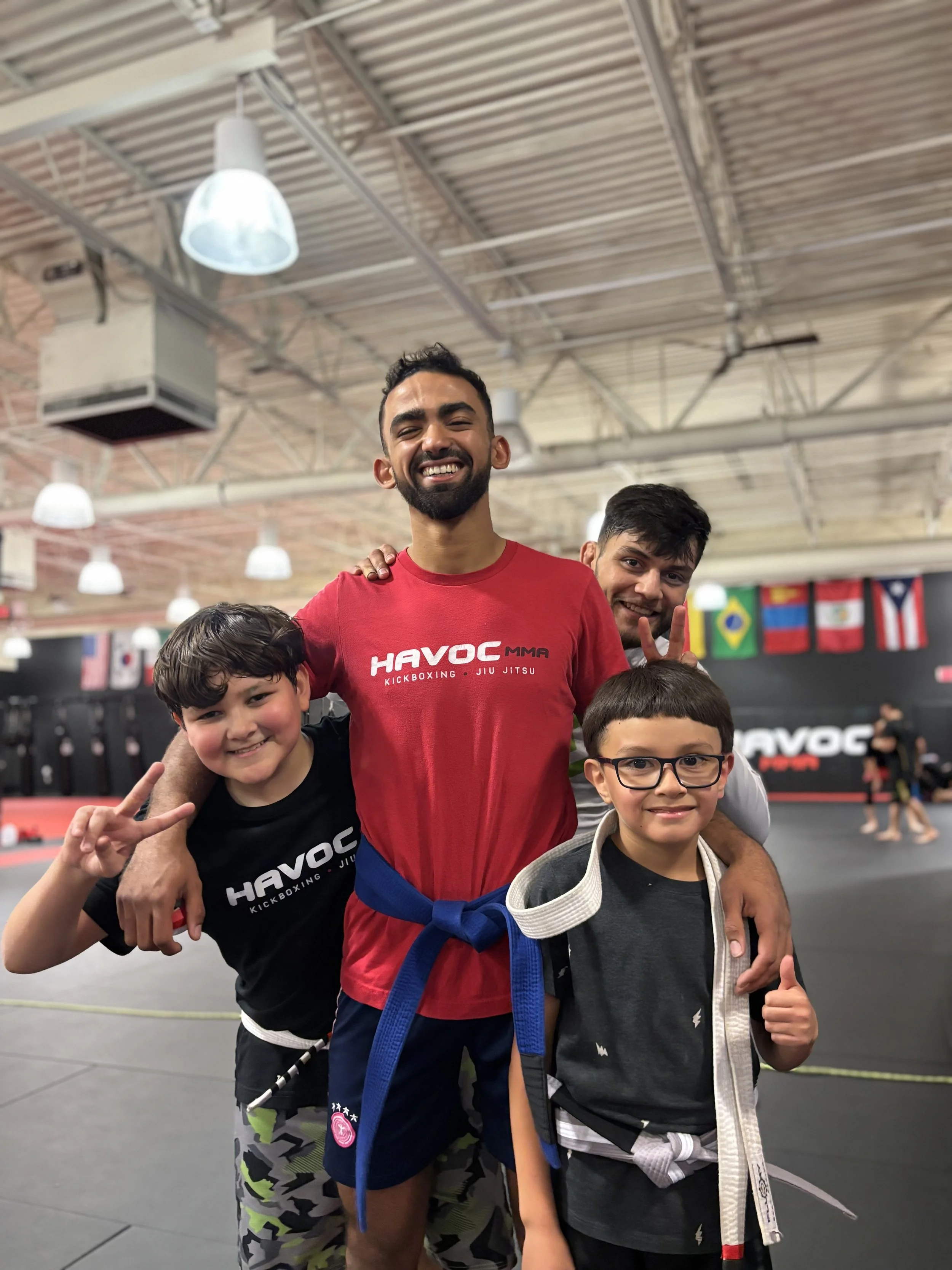Four people at a martial arts gym, smiling and posing for the photo. The two children are wearing martial arts uniforms and belts, while the two adults are in casual workout attire. The background shows martial arts flags and equipment.