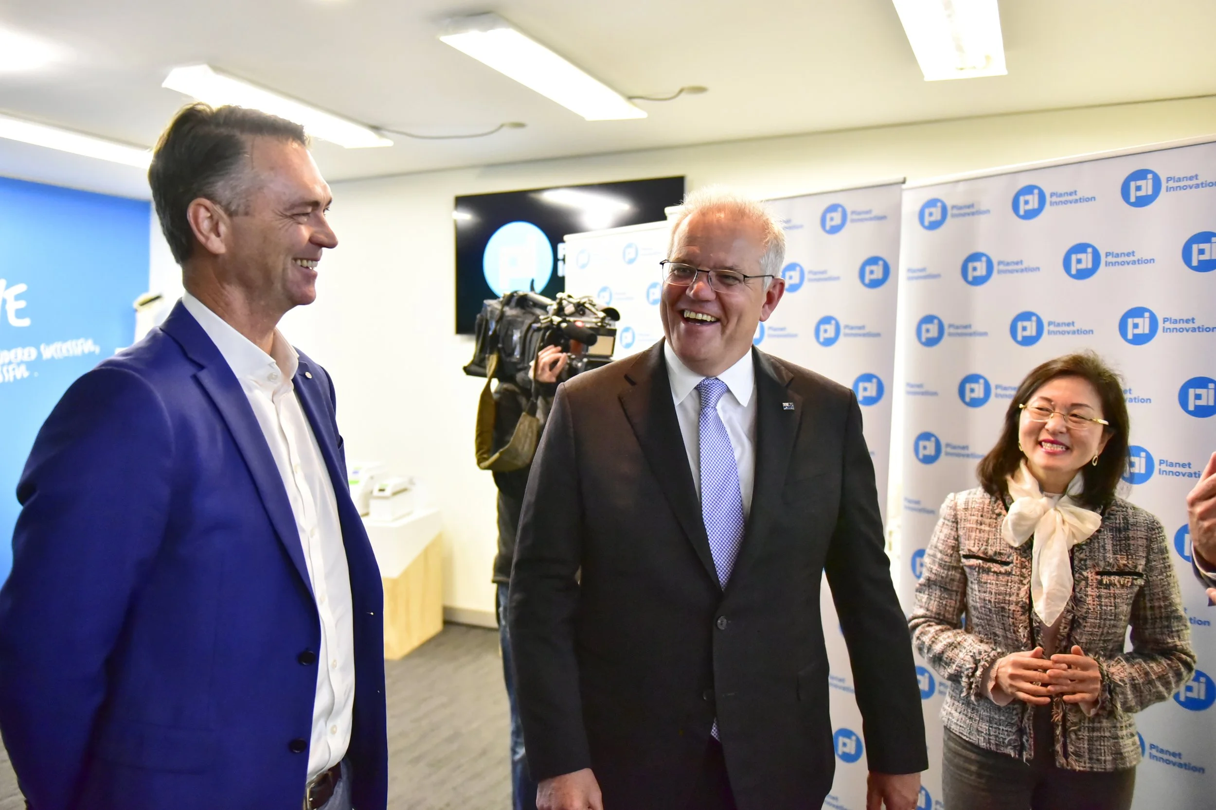 Three people, two men and a woman, are smiling and conversing at an event. They are dressed in business attire; the men are in suits, and the woman is wearing a patterned blazer with a scarf. There is a logo in the background that says "Planet Innova
