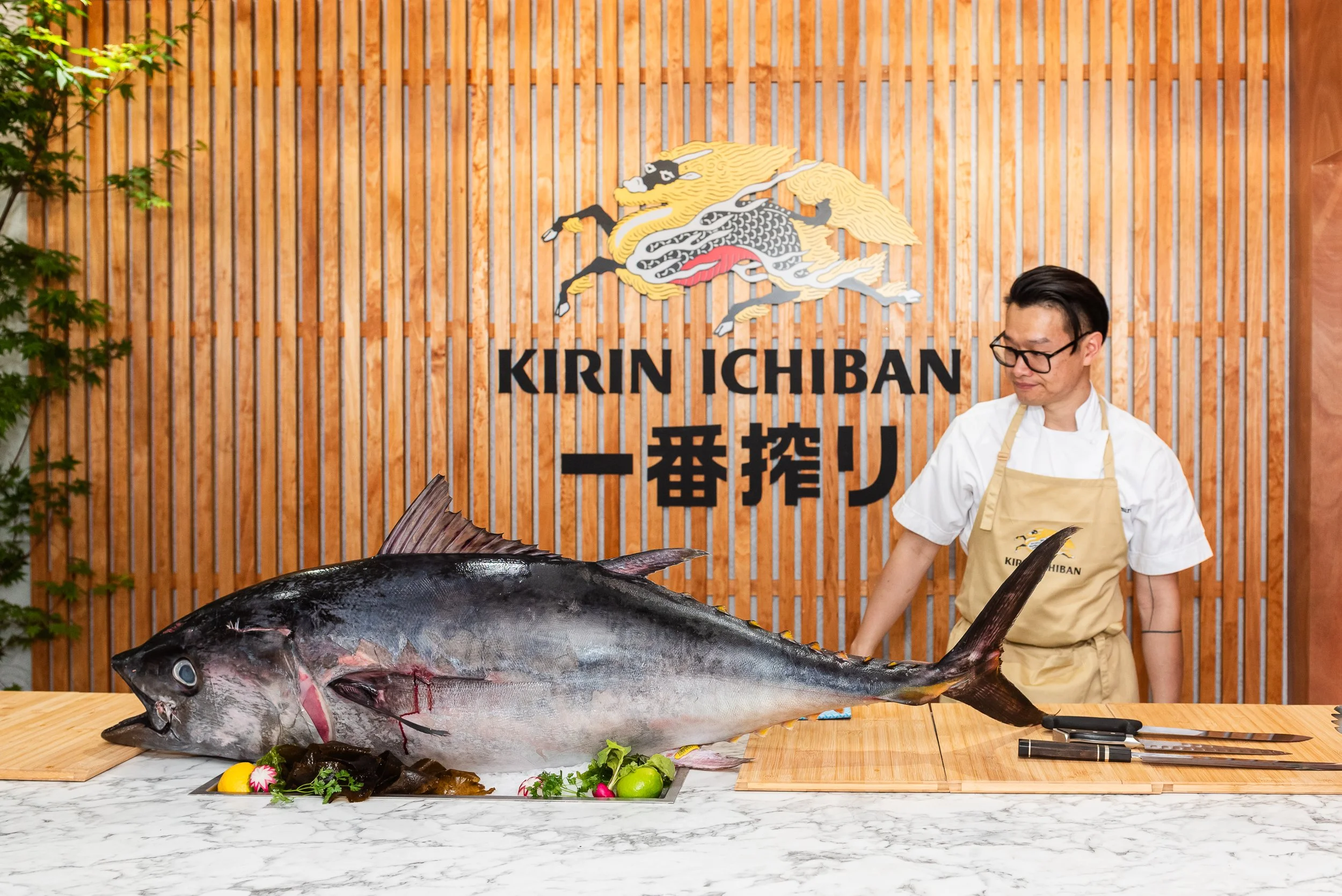 A chef presents a large fish on a marble countertop in a restaurant with a wooden wall background featuring the logo 'KIRIN ICHIBAN' and Japanese characters. The fish is on a bed of greens, lemon, and radish, with knives nearby.