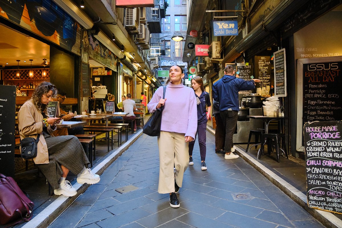 People walking and sitting outside cafes along a narrow street in an urban area.