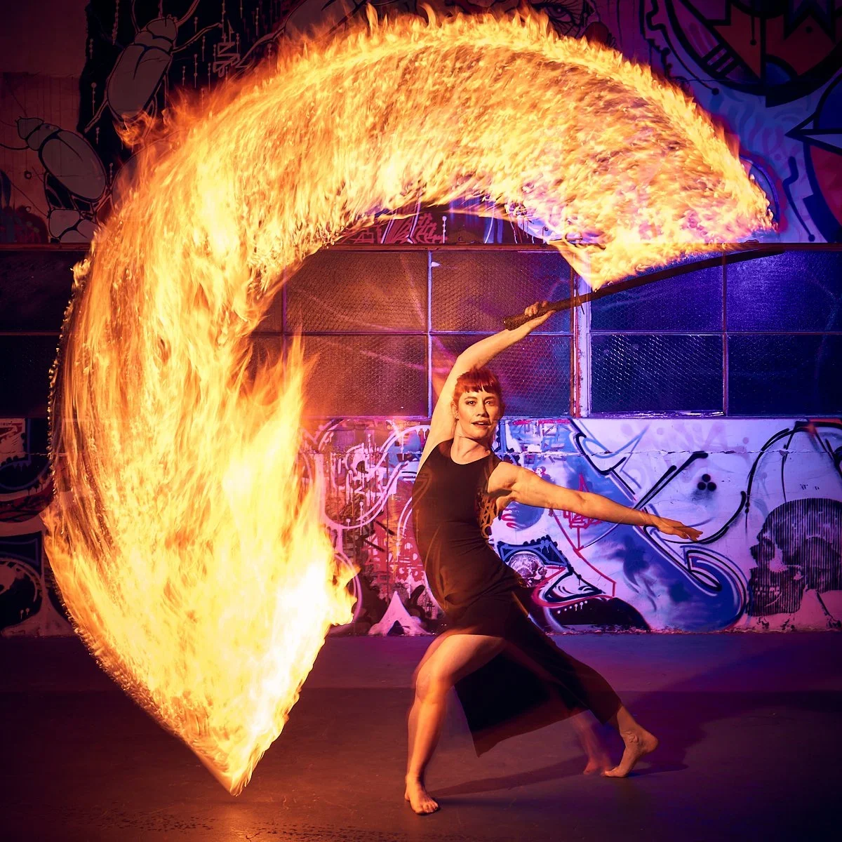 A female fire performer in a black dress spinning a flaming baton, creating a large arc of fire behind her in an urban, graffiti-covered setting.