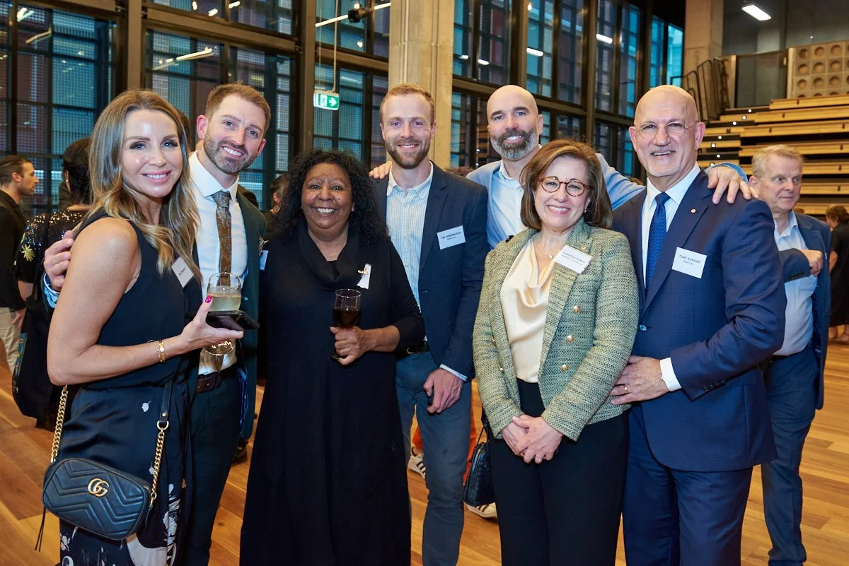Group of seven professionally dressed people at a social event, smiling and holding drinks, in a modern venue with large windows and wooden stairs in the background.