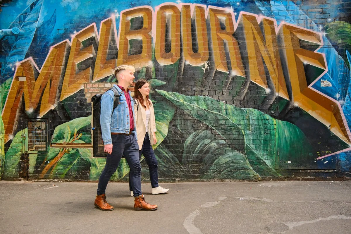 A mural with the word 'NEBRASKA' in large, colorful graffiti-style letters on a brick wall, featuring green leaves and blue tones in the background. Two young adults walk past it on the sidewalk.