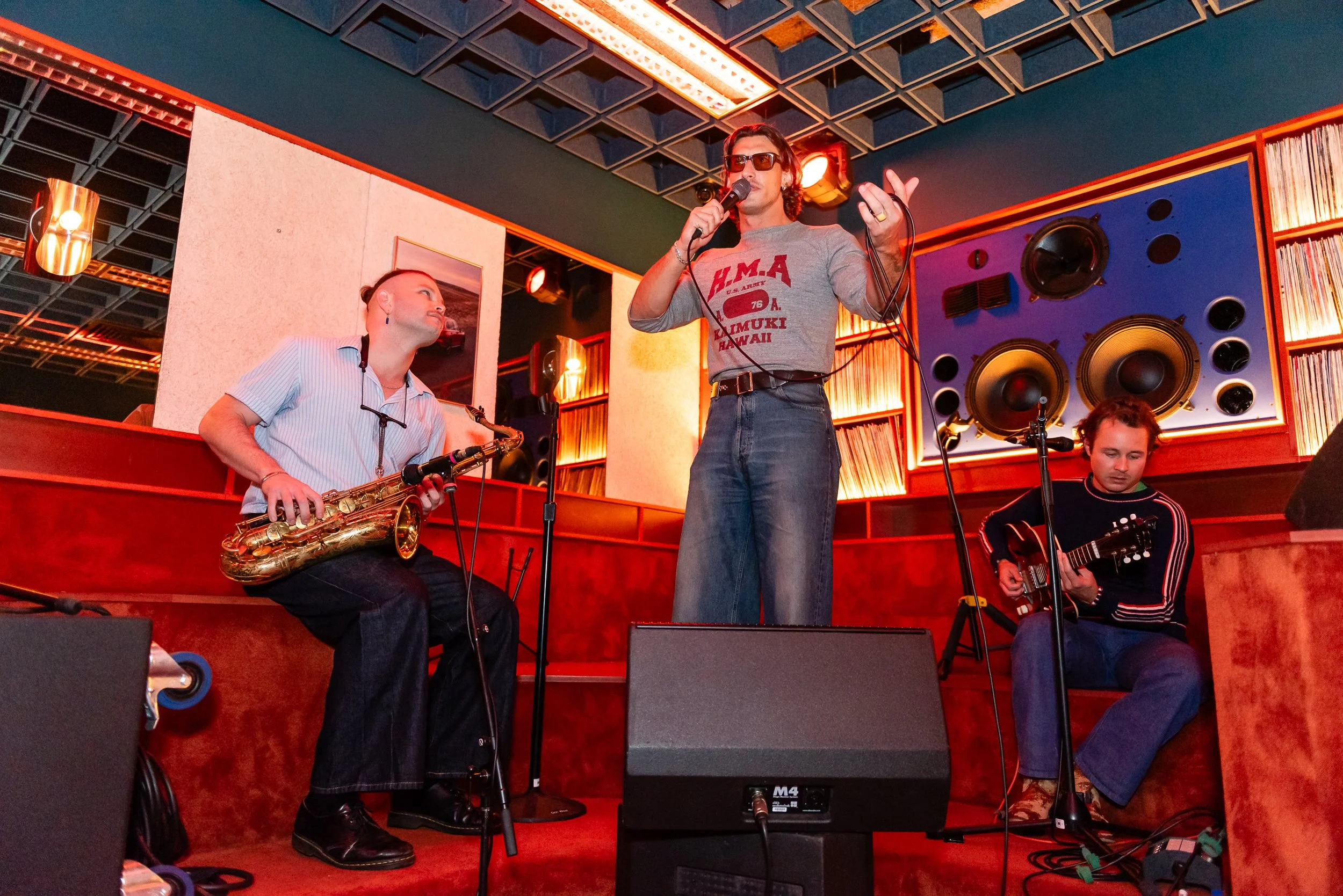 Four musicians performing live on a stage with orange lighting and acoustic panels. One woman with sunglasses and a microphone stands center, while a saxophonist and guitarist sit nearby.