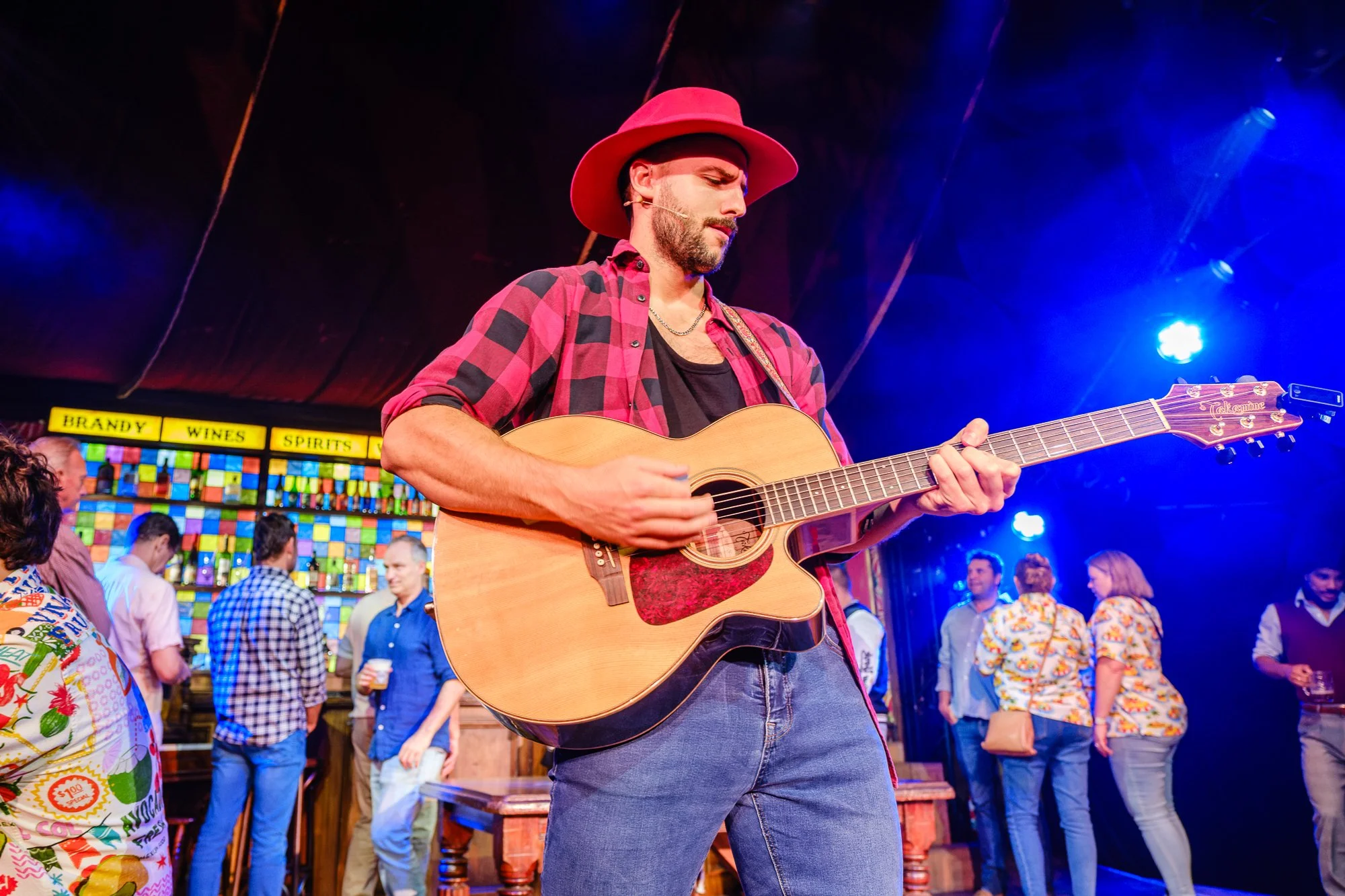 A man with a beard and mustache playing an acoustic guitar on stage at a lively event, wearing a pink hat, red and black checkered shirt, and dark pants, with a colorful crowd and a brightly lit background.