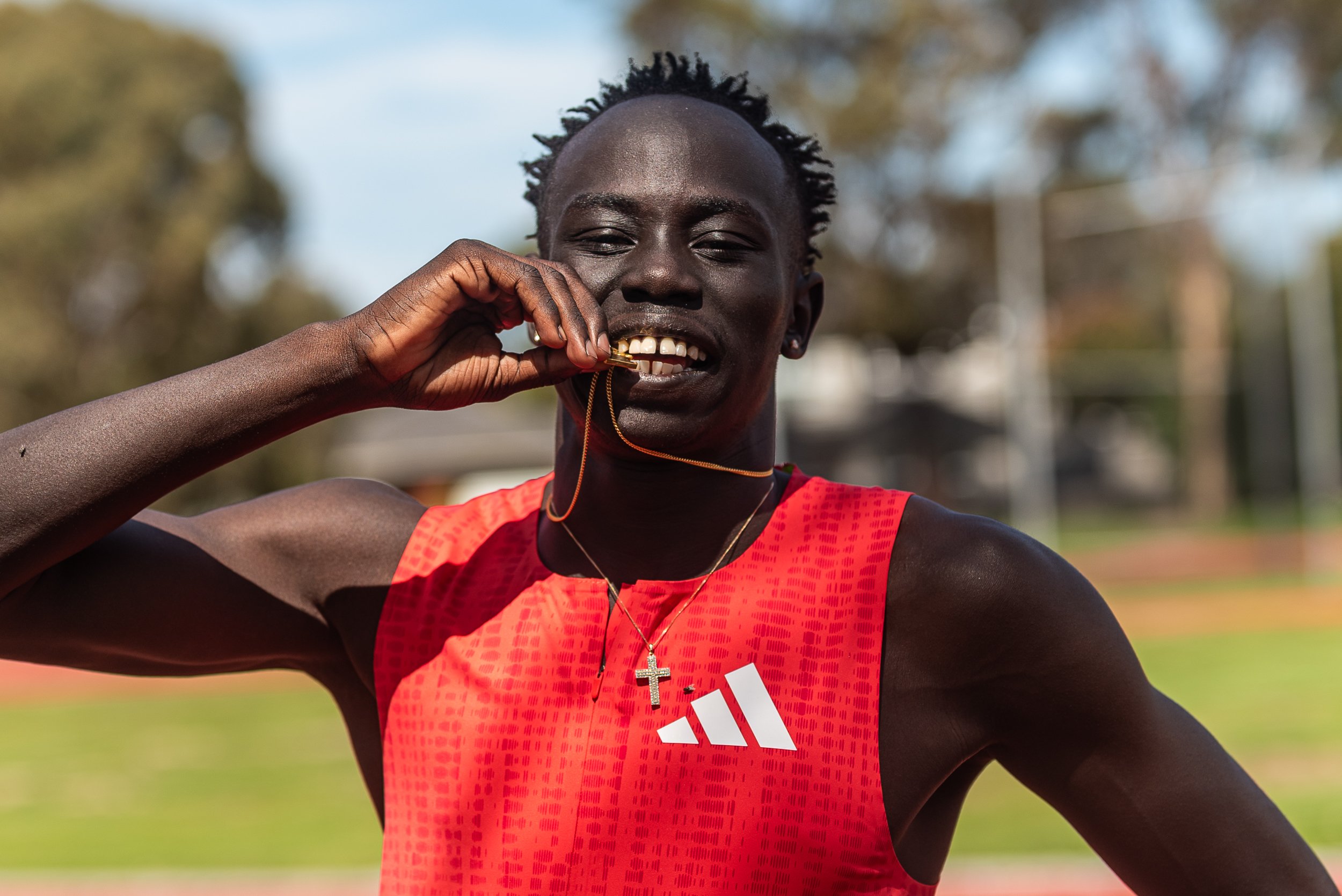 A male track athlete in a red Adidas sports top holding a gold medal with a chain near his mouth.