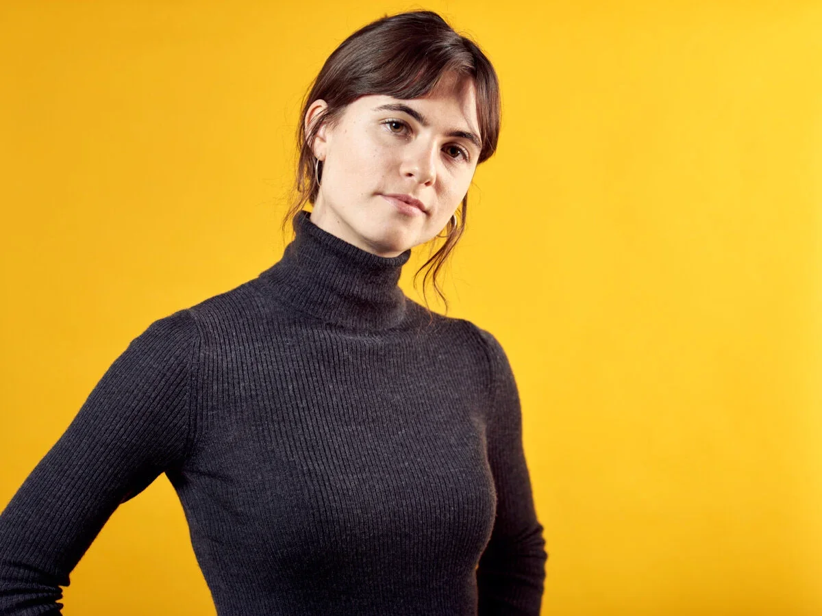 A woman with brown hair and fair skin wearing a black turtleneck sweater against a yellow background.