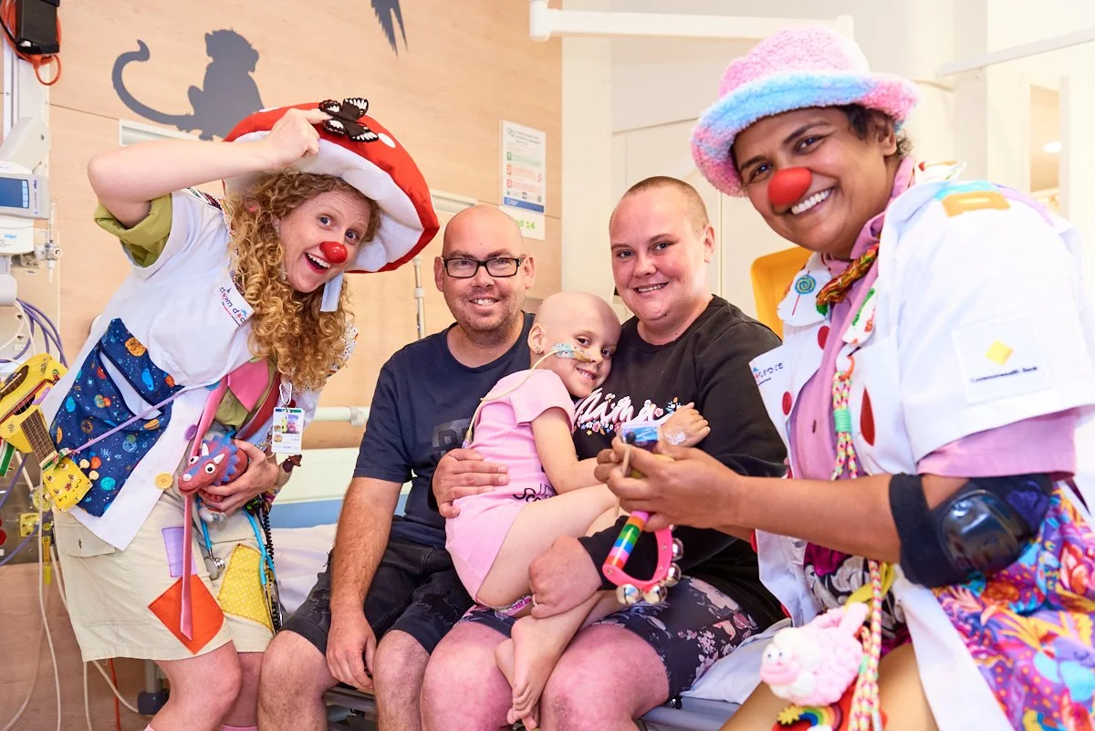 Medical staff and patients in a hospital room, with two children dressed as clowns. They are smiling, and the scene is cheerful and supportive.